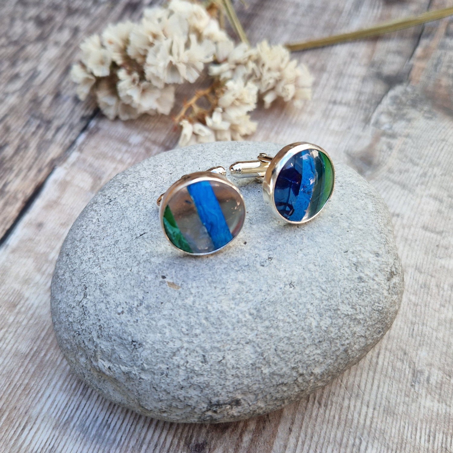 Cufflinks shown displayed on a grey stone with dried flowers in the background.