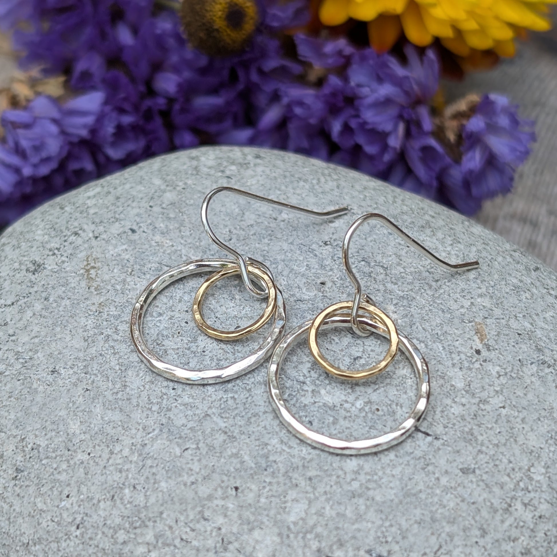 A pair of handmade earrings featuring two circles, one in silver and the other in gold, displayed on a smooth stone surface. The background includes a soft blur of purple and yellow flowers, enhancing the delicate craftsmanship of the earrings.