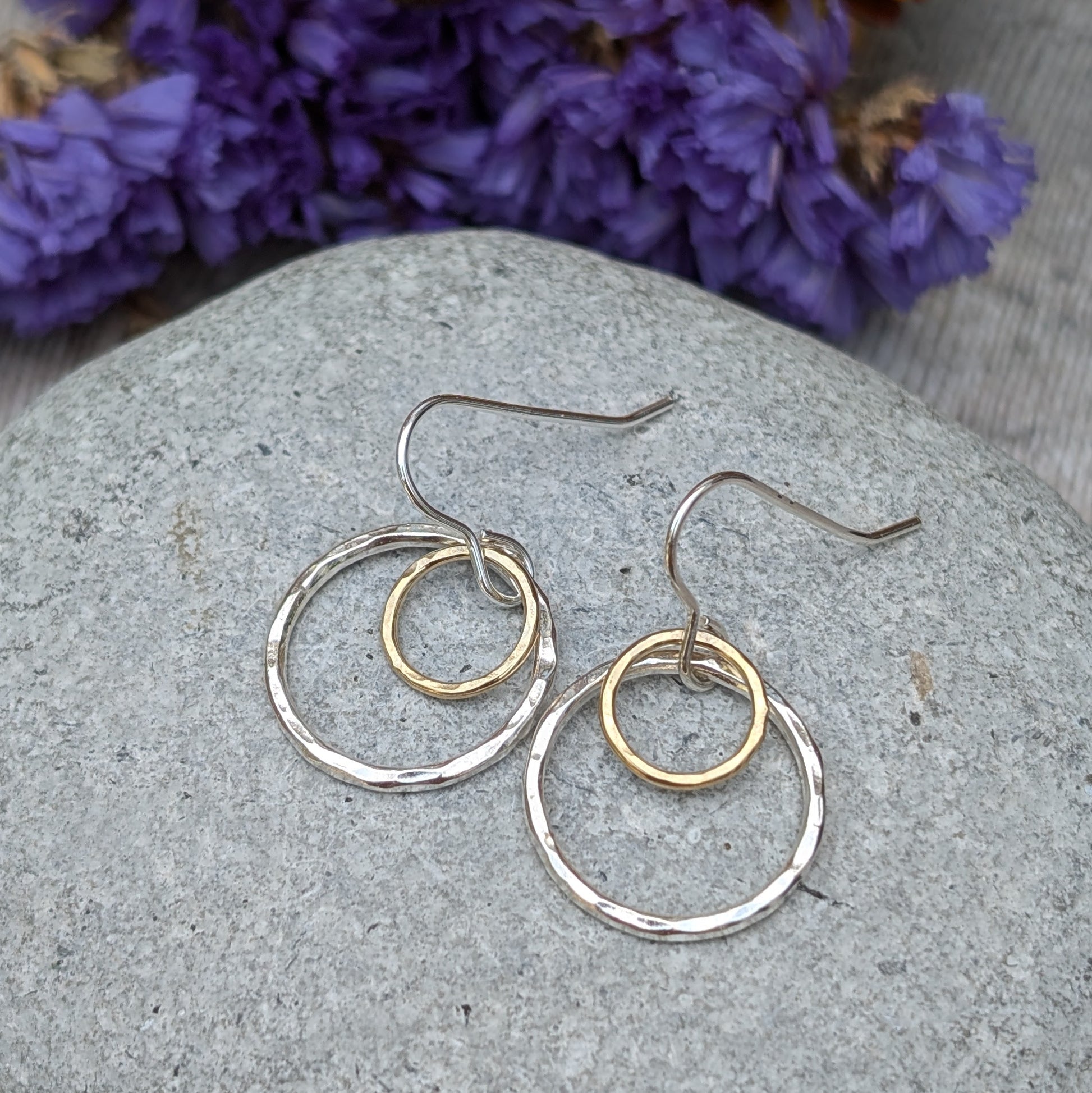 A pair of handmade earrings featuring two circles, one in silver and the other in gold, displayed on a smooth stone surface. The background includes a soft blur of purple and yellow flowers, enhancing the delicate craftsmanship of the earrings.