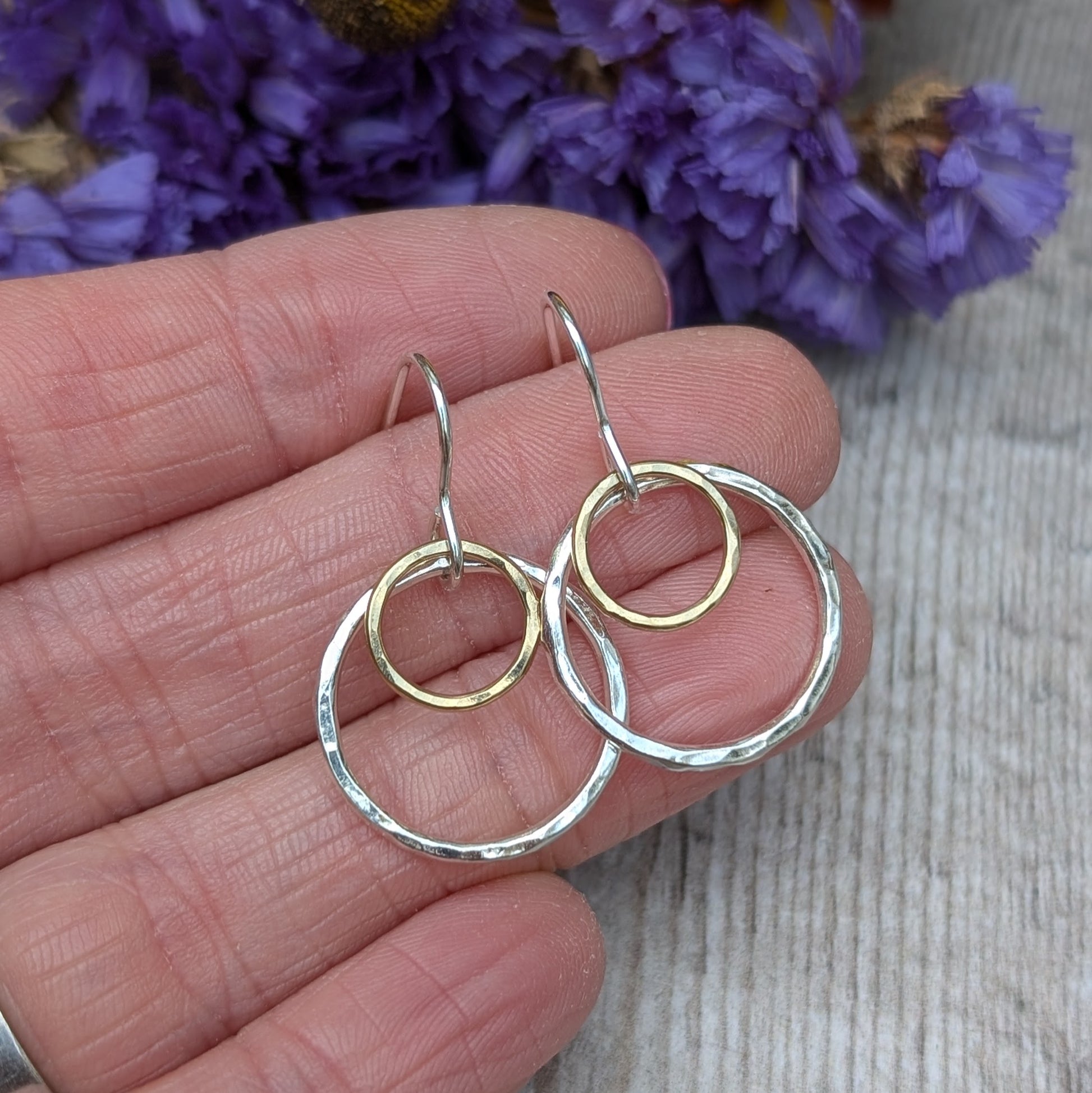 Close-up view of a hand holding a pair of handcrafted earrings with two hammered circles, one silver and one gold. The background shows a hint of purple flowers, adding a natural, rustic charm to the image.