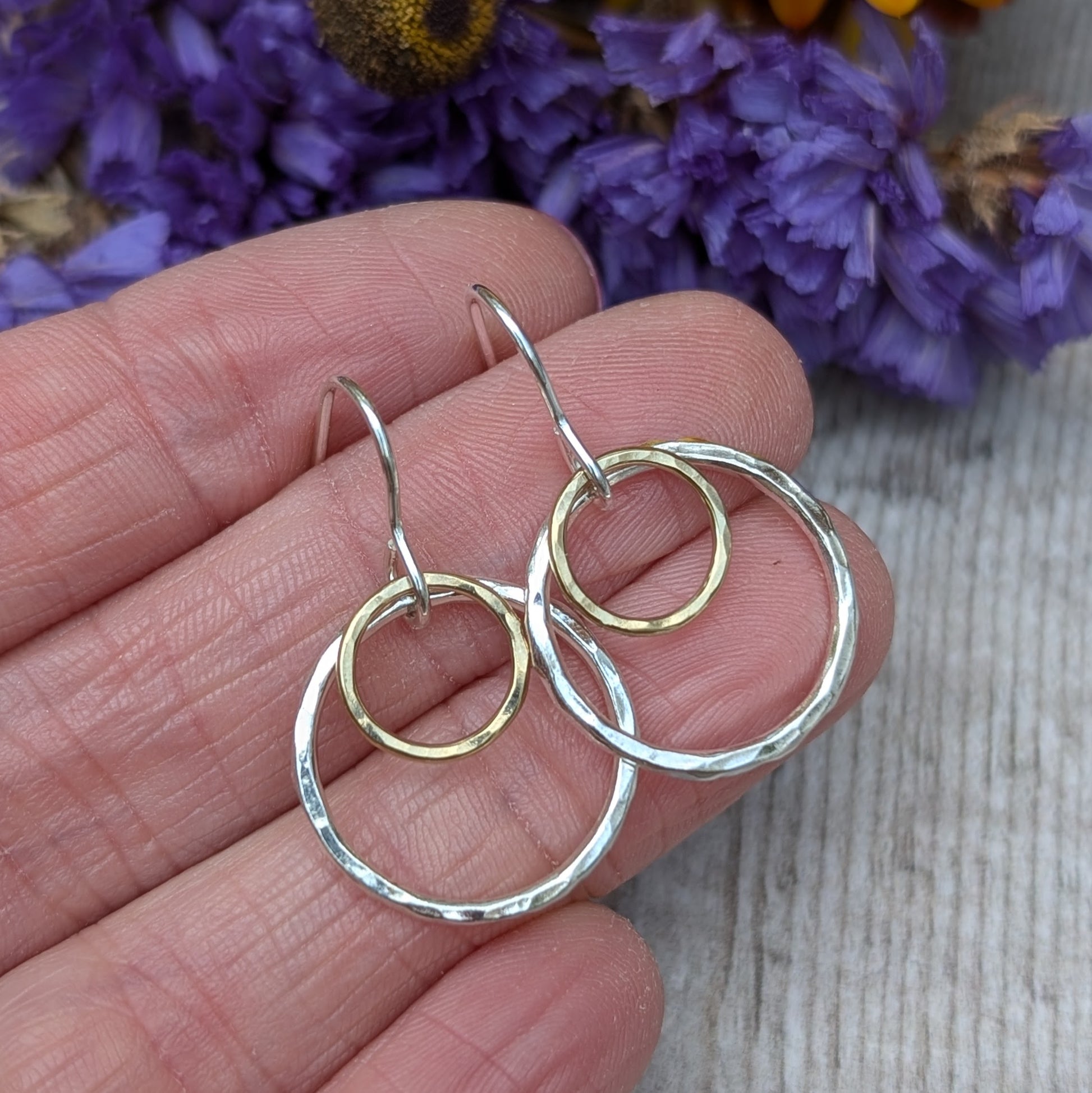 Close-up view of a hand holding a pair of handcrafted earrings with two hammered circles, one silver and one gold. The background shows a hint of purple flowers, adding a natural, rustic charm to the image.