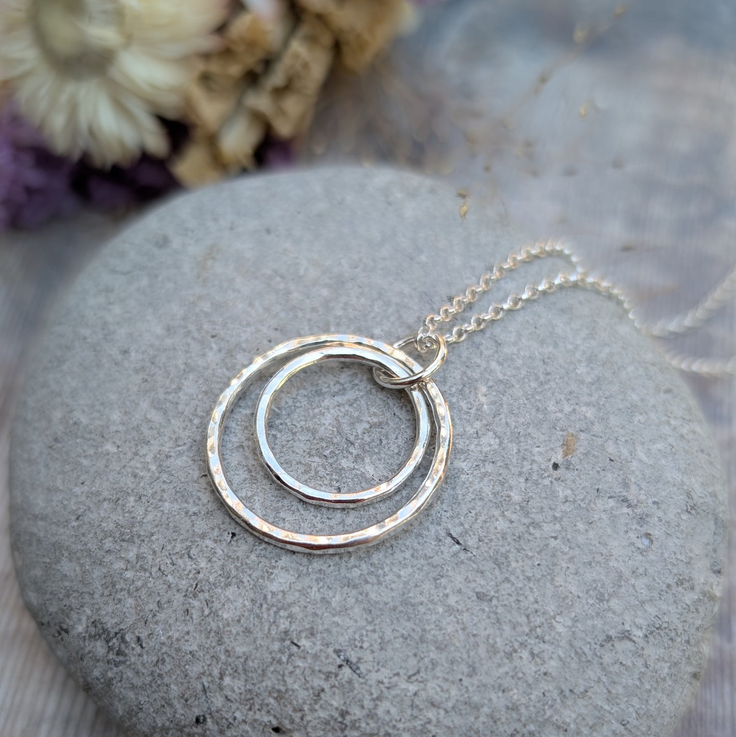 A handmade silver necklace with two interlinked hammered circles resting on a smooth grey stone. The necklace features a delicate silver chain and is styled with soft-focus dried flowers in the background.