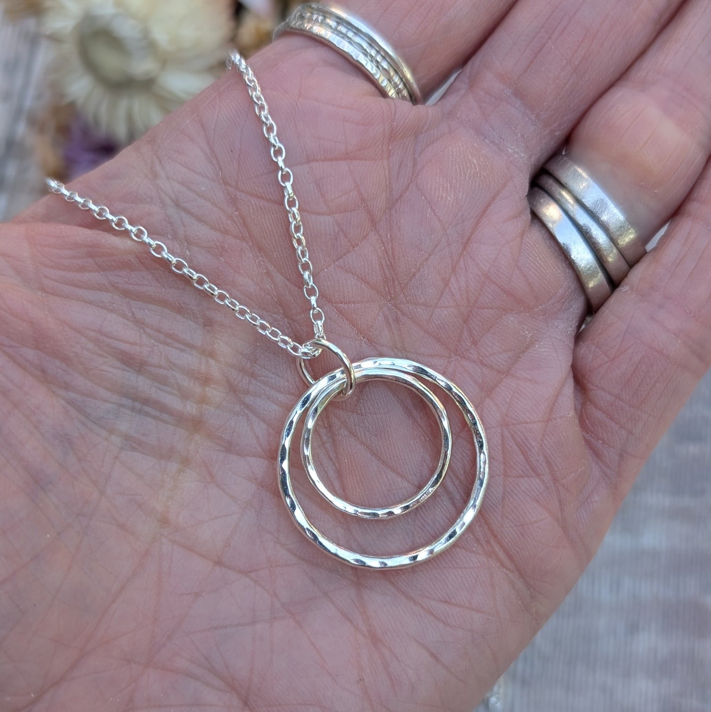 A close-up of a hand holding a handmade silver necklace with two hammered interlinked circles. The silver chain is looped through the top circle, and the hand is adorned with multiple silver rings.