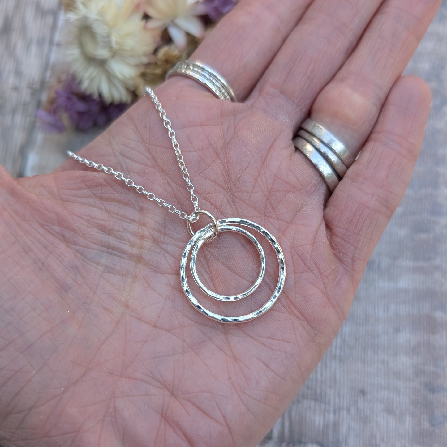 A close-up of a hand holding a handmade silver necklace with two hammered interlinked circles. The silver chain is looped through the top circle, and the hand is adorned with multiple silver rings.