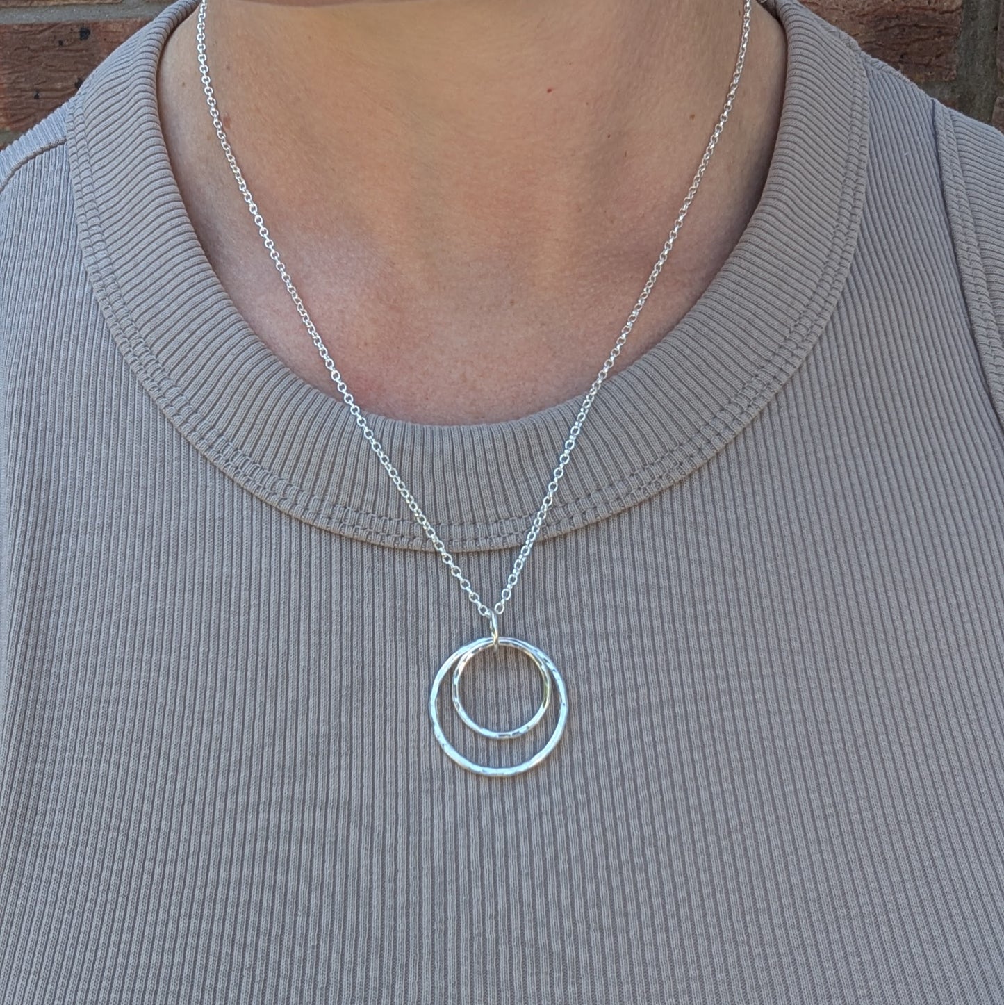 A handmade silver double circle necklace worn on a person’s neck. The two hammered silver circles hang from a fine silver chain, shown against a ribbed taupe top.