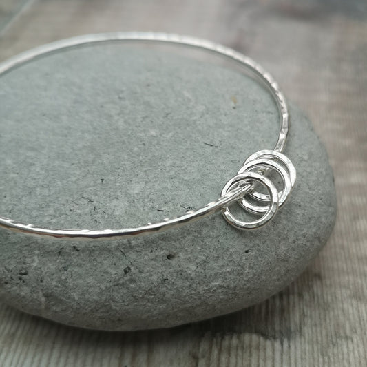 A close-up of a sterling silver bangle with a hammered finish, adorned with three small hammered rings. The minimalist design is highlighted by the polished surface and the texture of the rings.