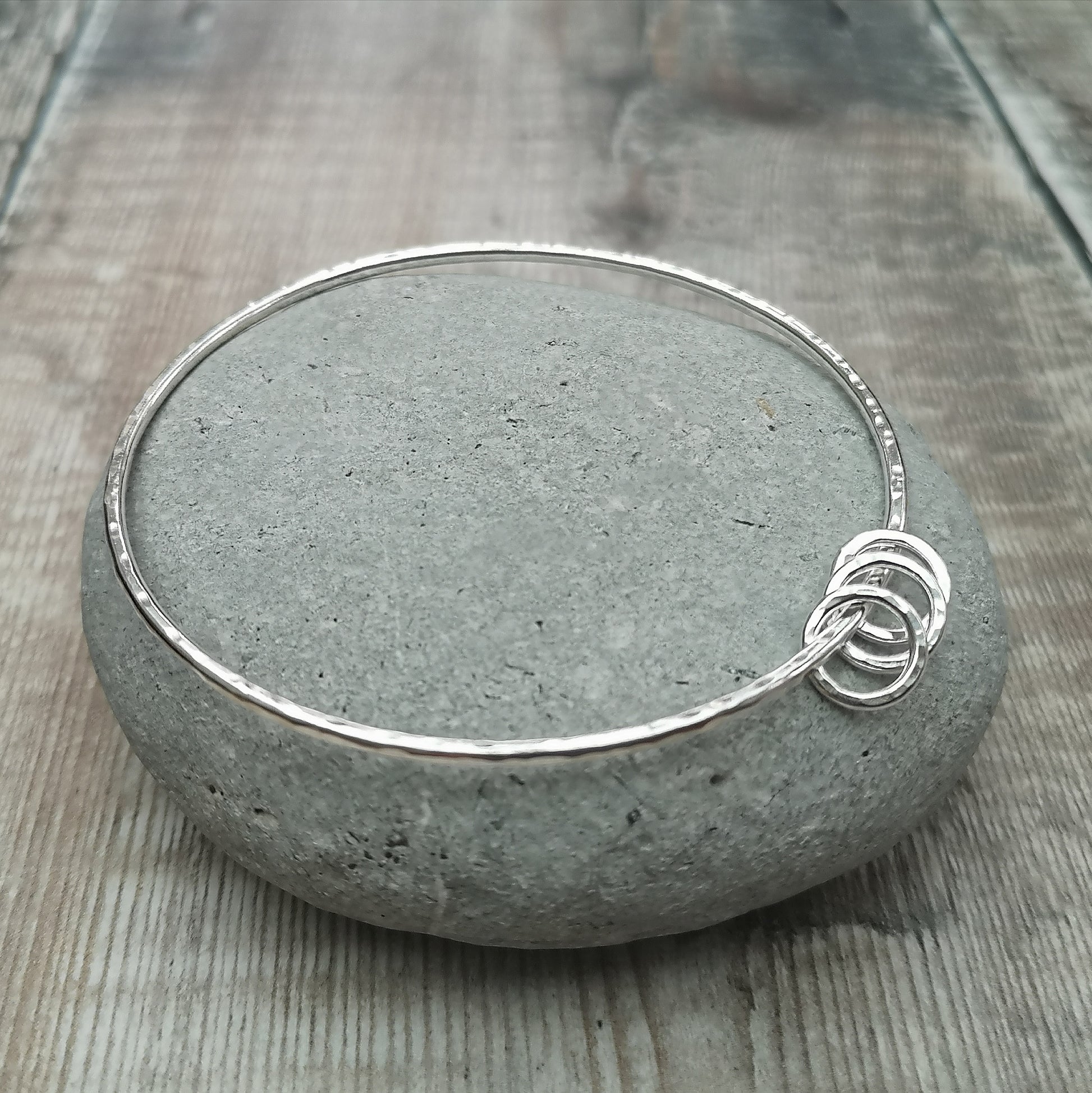 A close-up of a sterling silver bangle with a hammered finish, adorned with three small hammered rings. The minimalist design is highlighted by the polished surface and the texture of the rings.