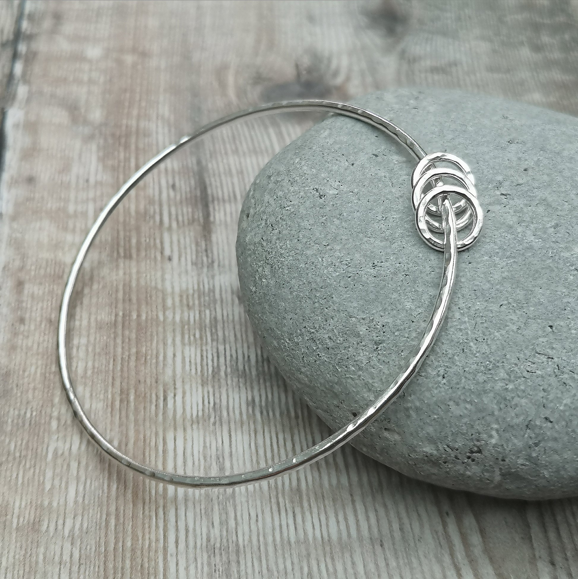 A sterling silver hammered bangle with three small rings displayed on a smooth grey stone. The sleek, simple design of the bangle and rings creates a modern and elegant look.