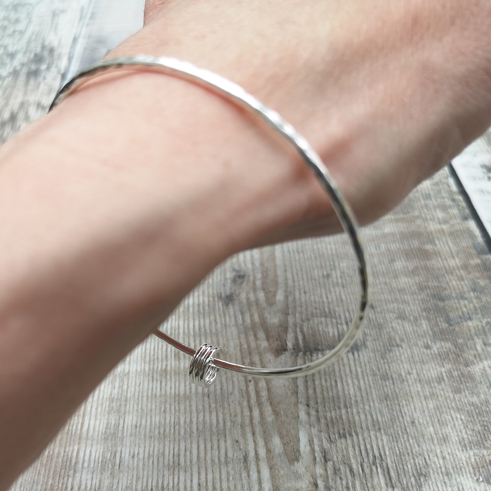 A sterling silver hammered bangle with three small rings worn on a wrist. The lightweight design and the textured surface of the bangle catch the light, showcasing its delicate and timeless style.