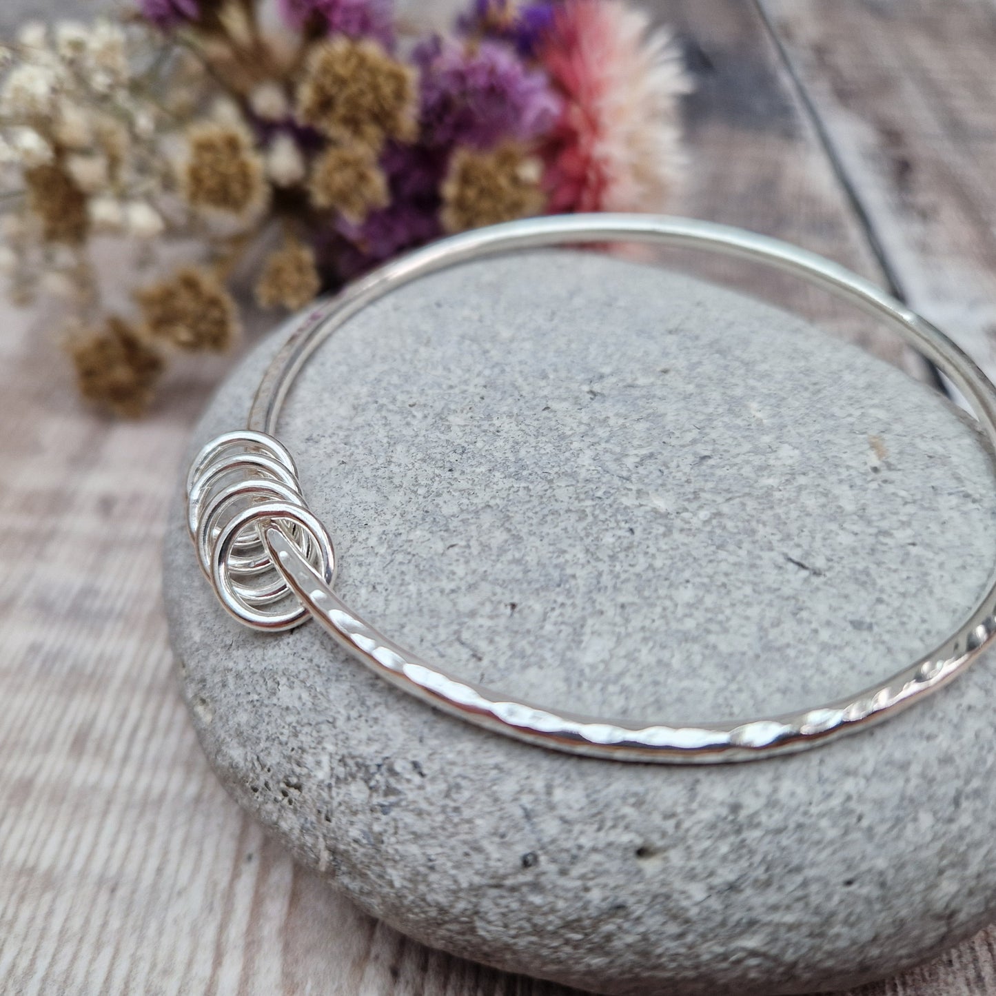 A handmade sterling silver bangle with four silver rings resting on its hammered surface, placed on a smooth stone with a background of soft, dried flowers in pink and cream tones.