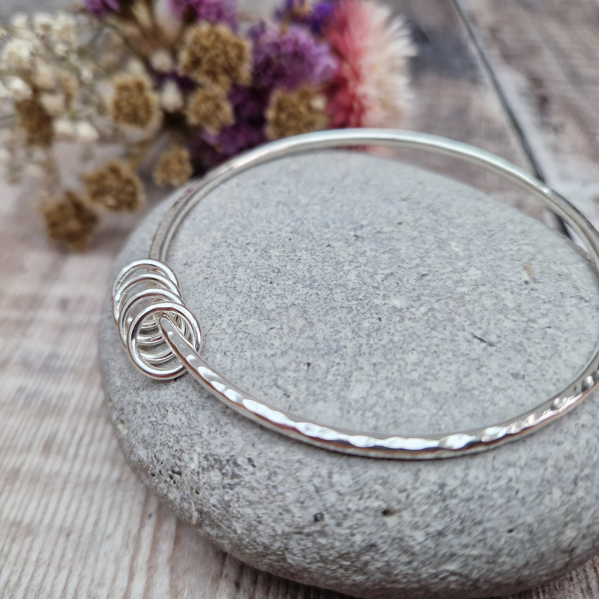 A handmade sterling silver bangle with four silver rings resting on its hammered surface, placed on a smooth stone with a background of soft, dried flowers in pink and cream tones.