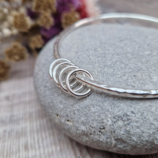 A handmade sterling silver bangle with four silver rings resting on its hammered surface, placed on a smooth stone with a background of soft, dried flowers in pink and cream tones.