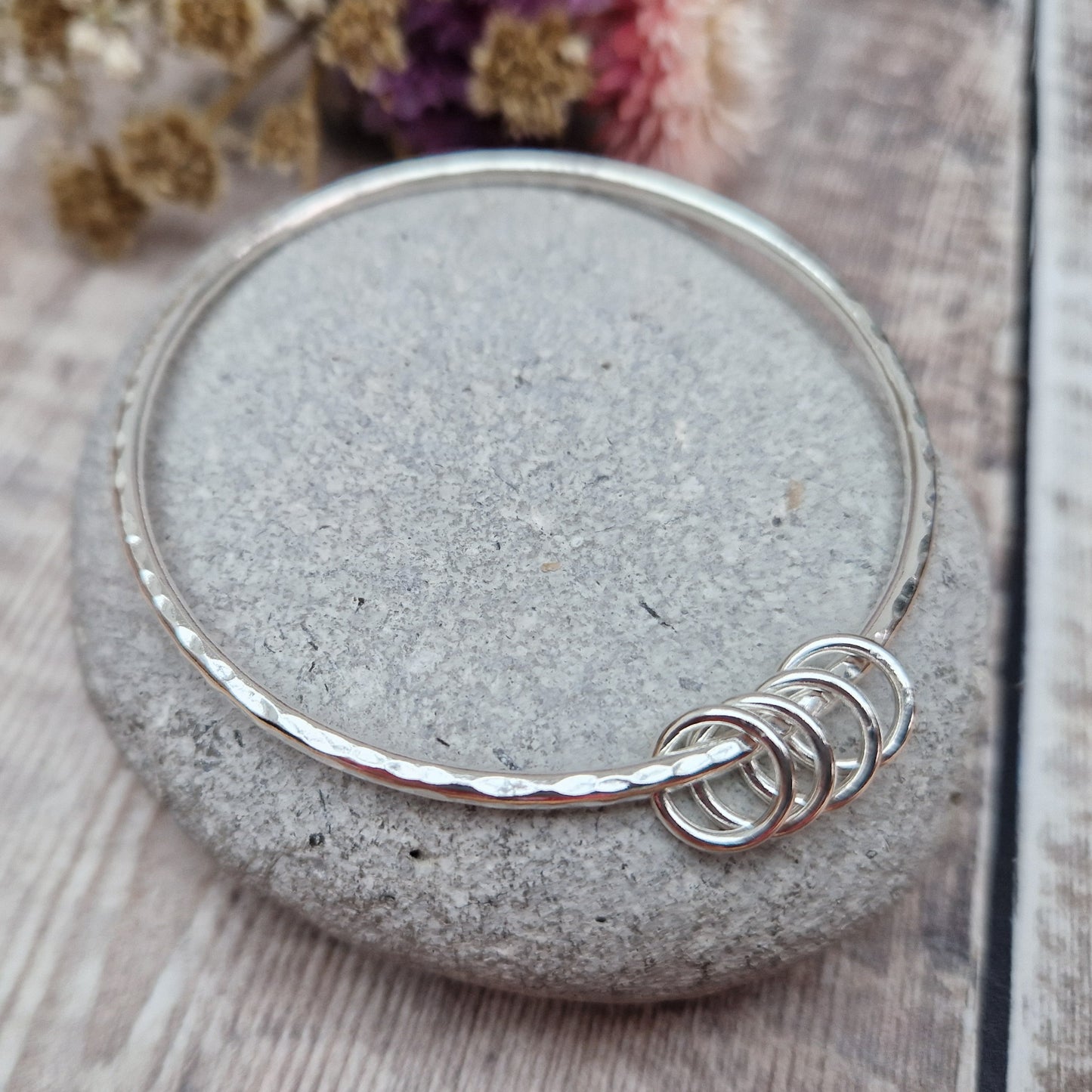 A handmade sterling silver bangle with four silver rings resting on its hammered surface, placed on a smooth stone with a background of soft, dried flowers in pink and cream tones.