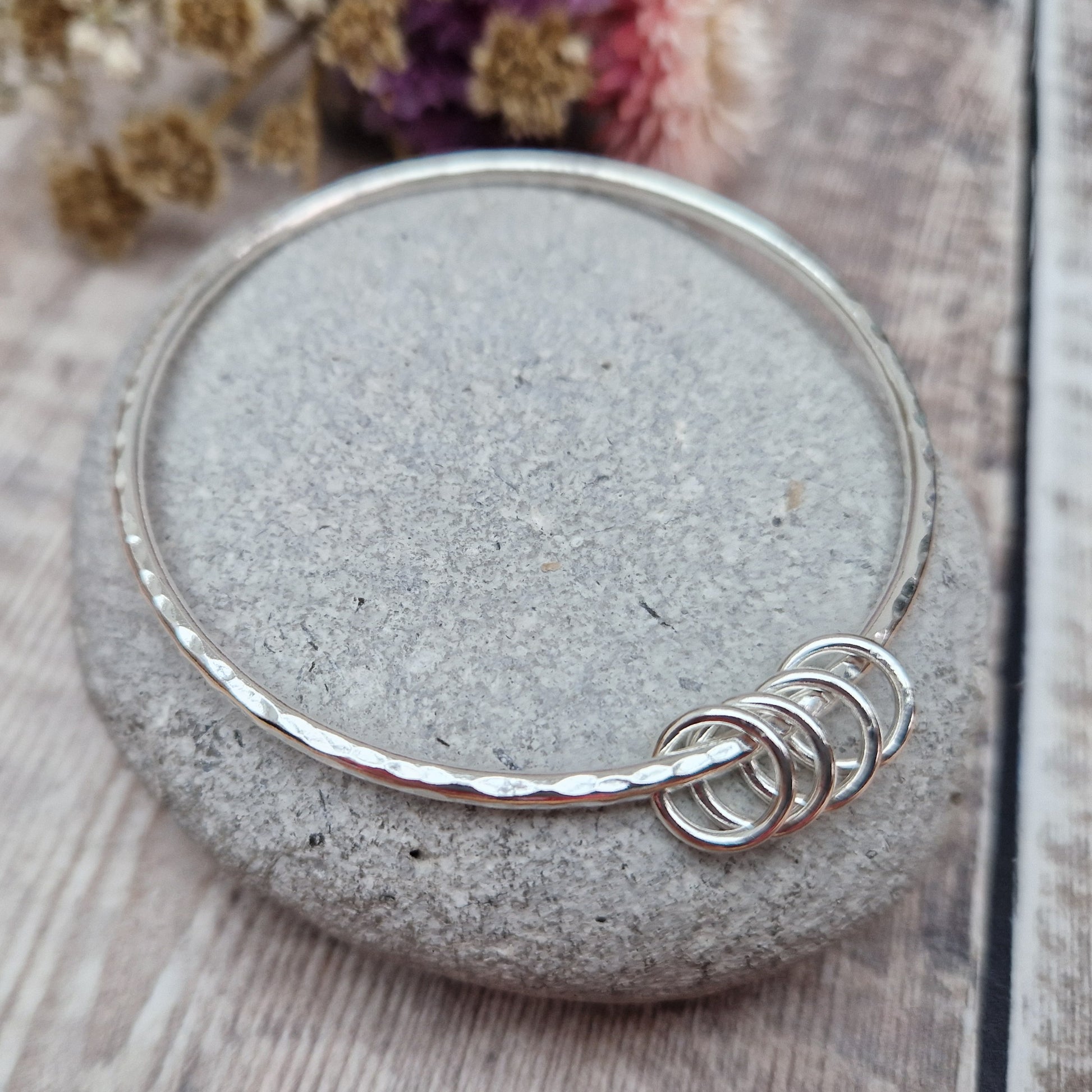 A handmade sterling silver bangle with four silver rings resting on its hammered surface, placed on a smooth stone with a background of soft, dried flowers in pink and cream tones.