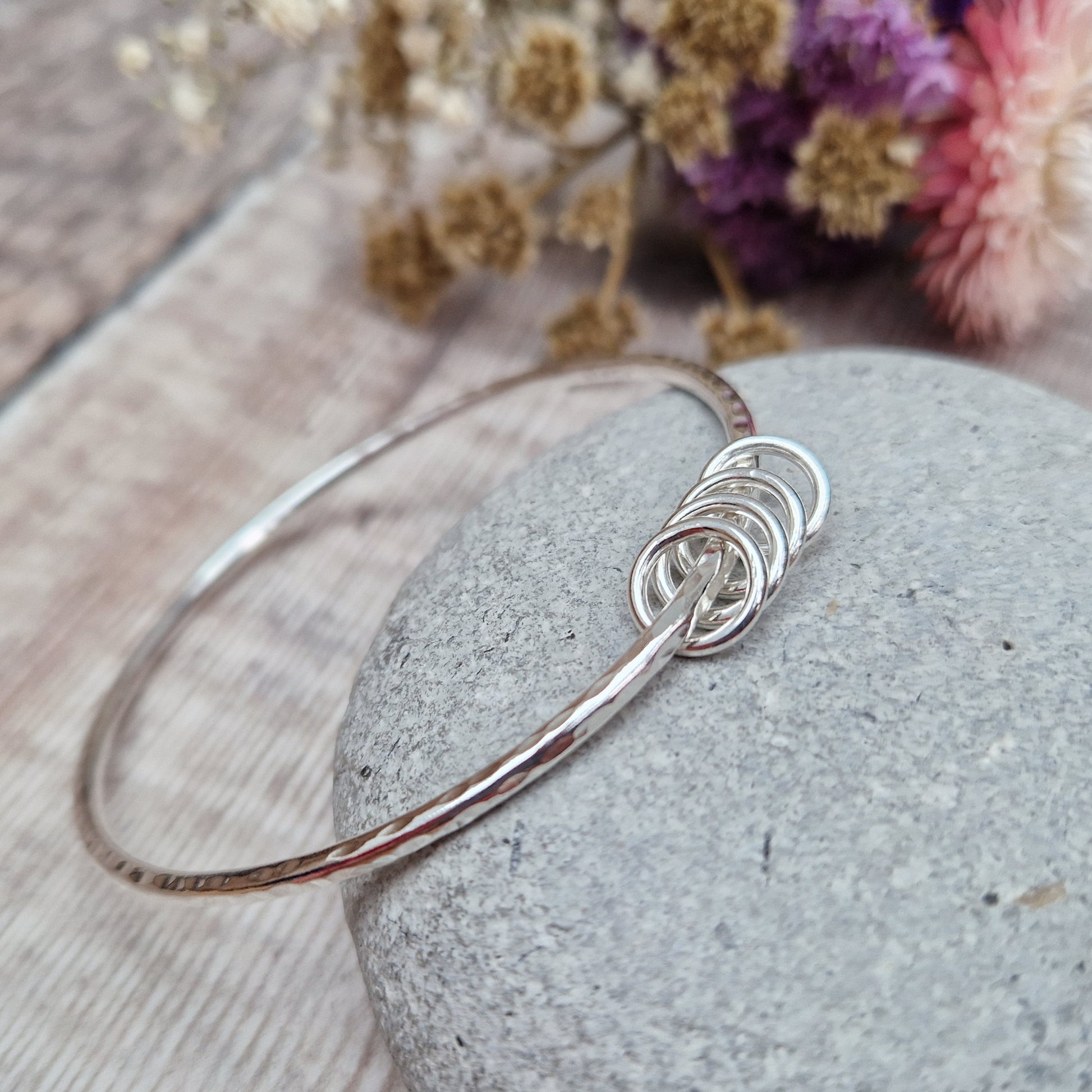 A handmade sterling silver bangle with four silver rings resting on its hammered surface, placed on a smooth stone with a background of soft, dried flowers in pink and cream tones.