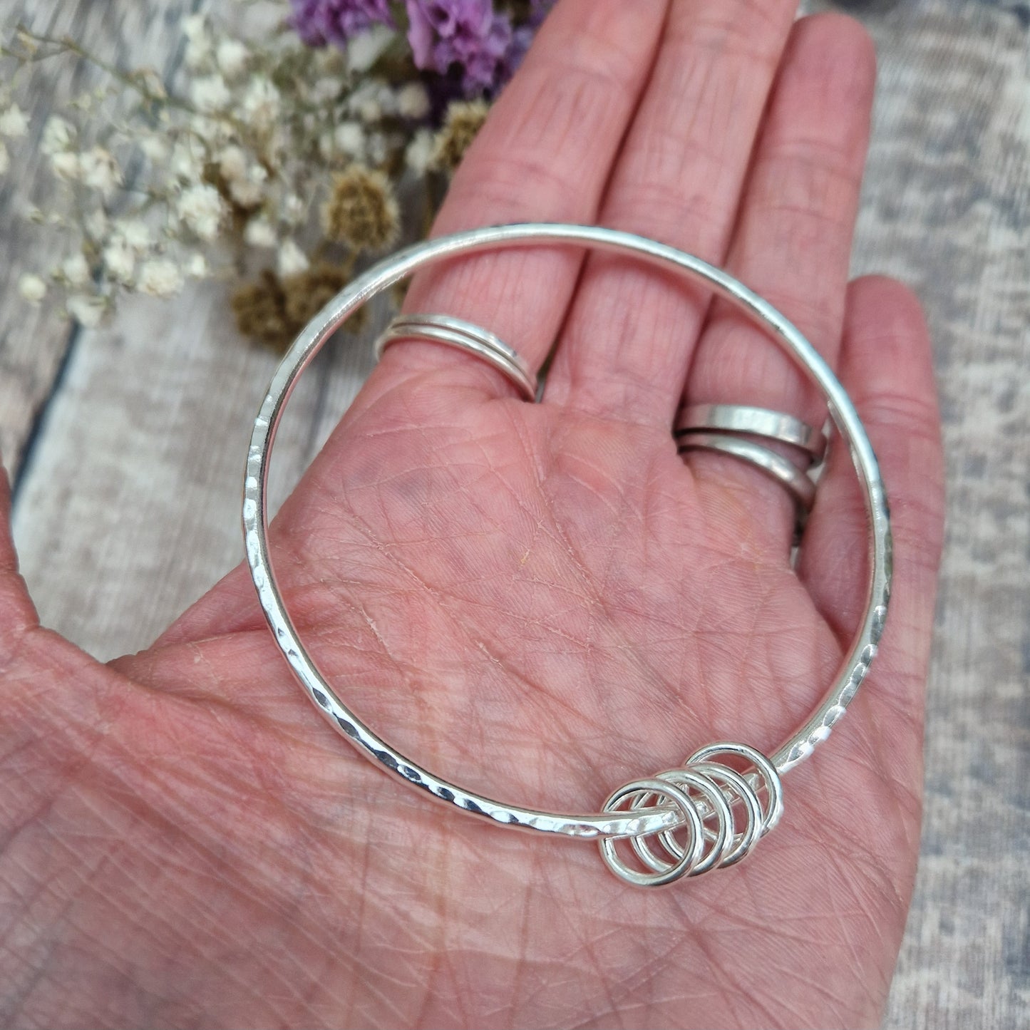 A close-up of a handmade sterling silver bangle with four small silver rings, shown in an open hand for scale. The bangle’s hammered texture shines under the light, highlighting its simplicity and charm.