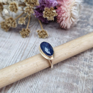 9ct gold ring with dark blue colour sapphire gemstone, oval shaped. Set in raised gold surround. Gemstone set lengthways across ring. Slight texture to stone, like diamond cut.