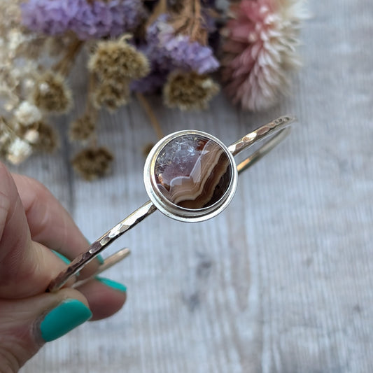 Close-up of a hand holding a sterling silver bangle with a hammered texture, featuring a striking Agua Nueva Agate stone with natural banding in shades of brown and cream, against a backdrop of dried flowers.