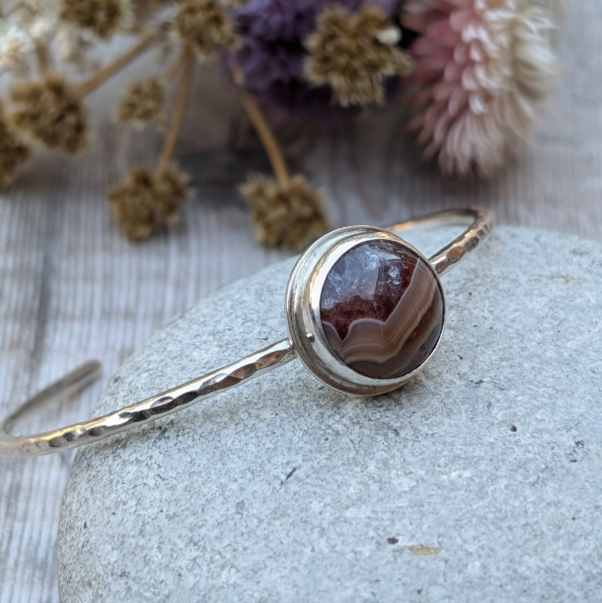 Close-up of a handmade sterling silver bangle with a hammered finish, featuring a striking Agua Nueva Agate stone with warm, natural banding, resting on a smooth grey stone with dried flowers in the background.