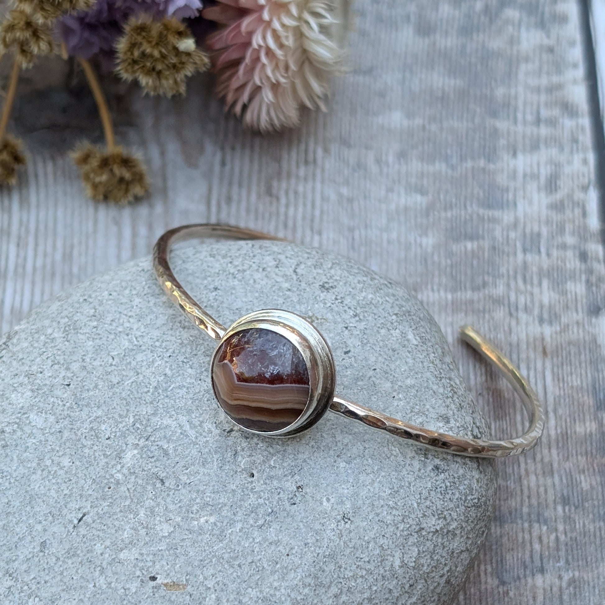 Close-up of a handmade sterling silver bangle with a hammered finish, featuring a striking Agua Nueva Agate stone with warm, natural banding, resting on a smooth grey stone with dried flowers in the background.