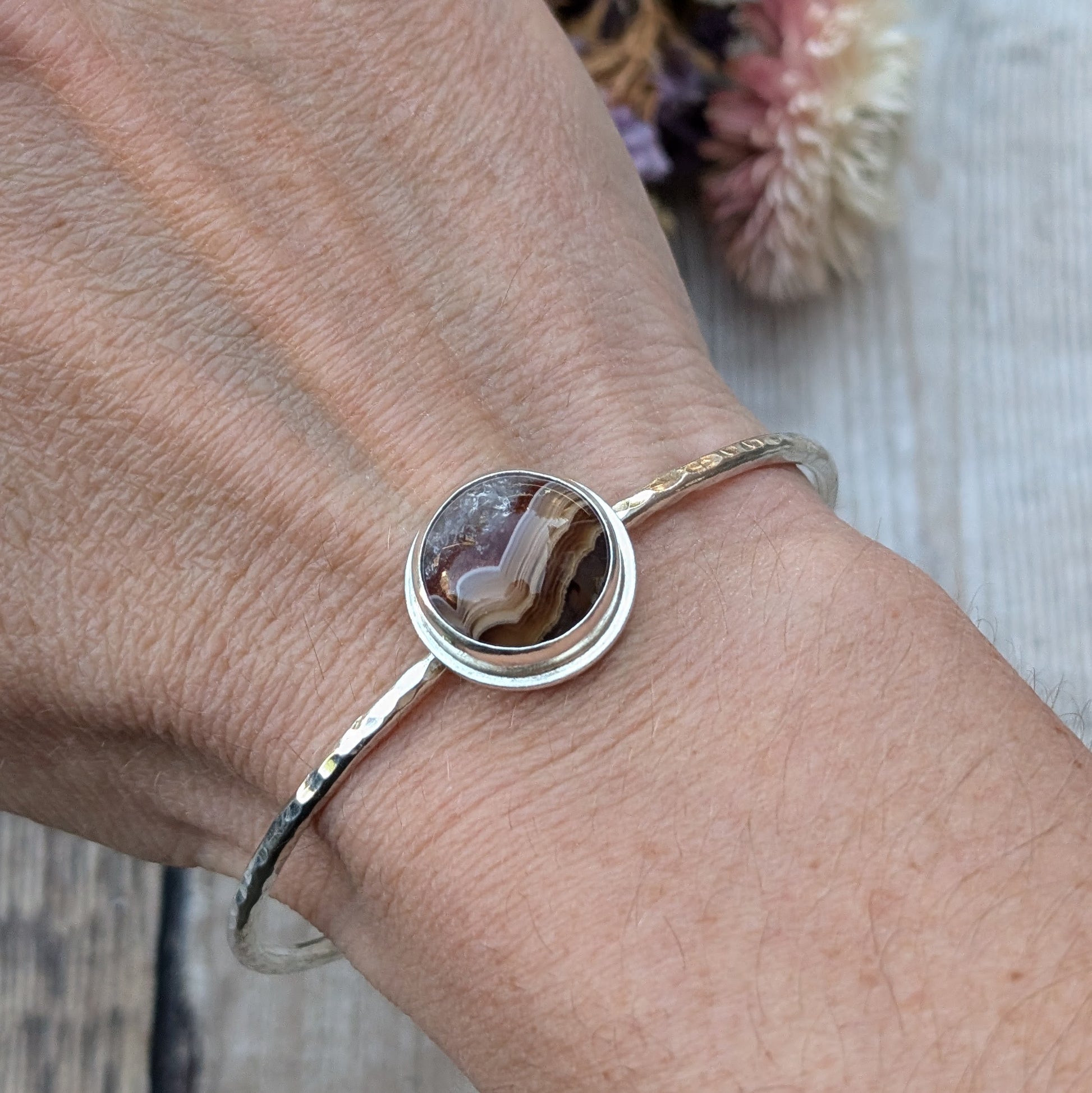 Close-up of a person's wrist wearing a handmade sterling silver bangle with a hammered finish, showcasing an Agua Nueva Agate stone with intricate layers of brown, white, and crystal formations.