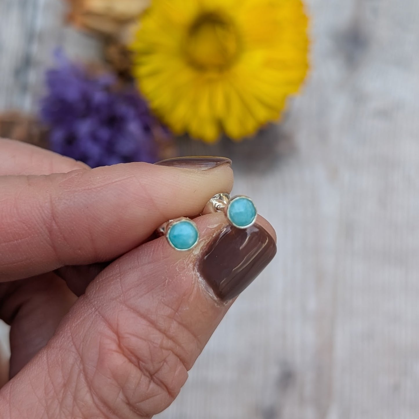 A close-up of fingers holding a pair of silver amazonite studs, highlighting the polished silver setting and blue-green gemstones with flowers softly out of focus in the background.