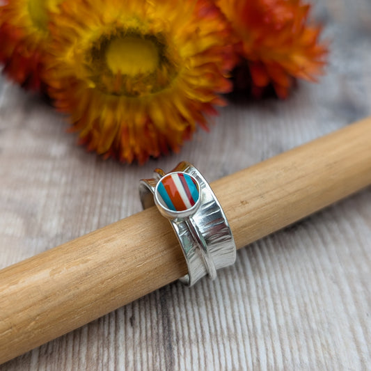 Bark textured silver spinner ring with a Surfite cabochon, displayed on a ring mandrel with orange flowers in the background.
