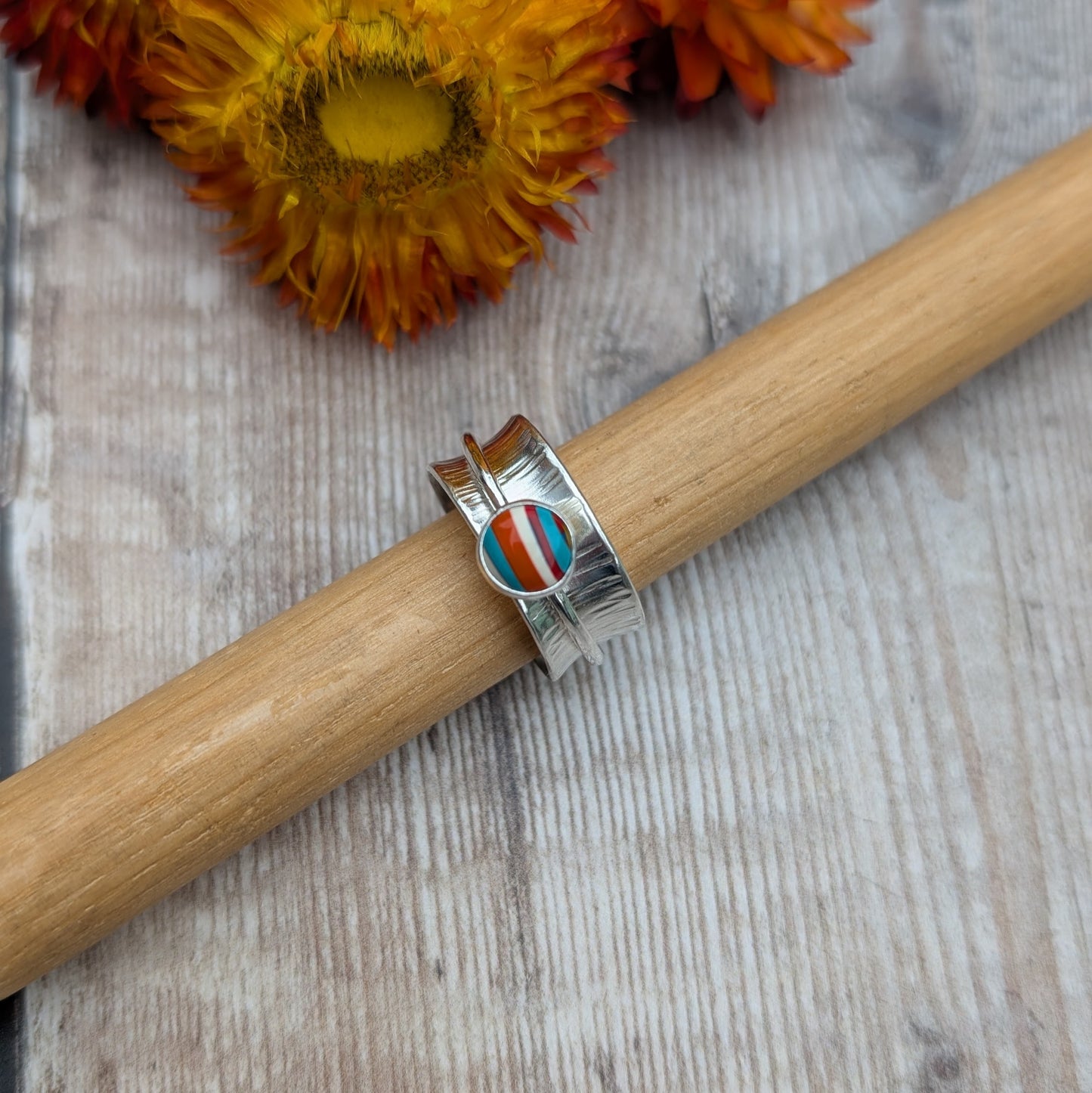 Bark textured silver spinner ring with a Surfite cabochon, displayed on a ring mandrel with orange flowers in the background.