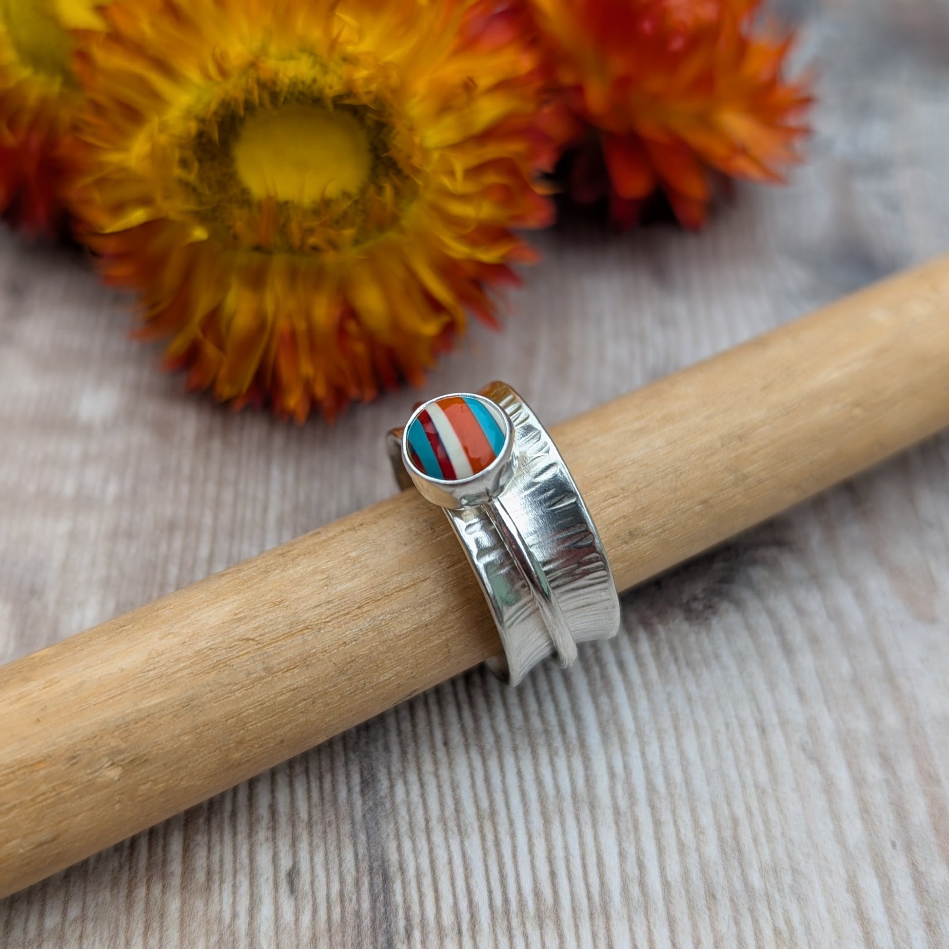 Bark textured silver spinner ring with a Surfite cabochon, displayed on a ring mandrel with orange flowers in the background.