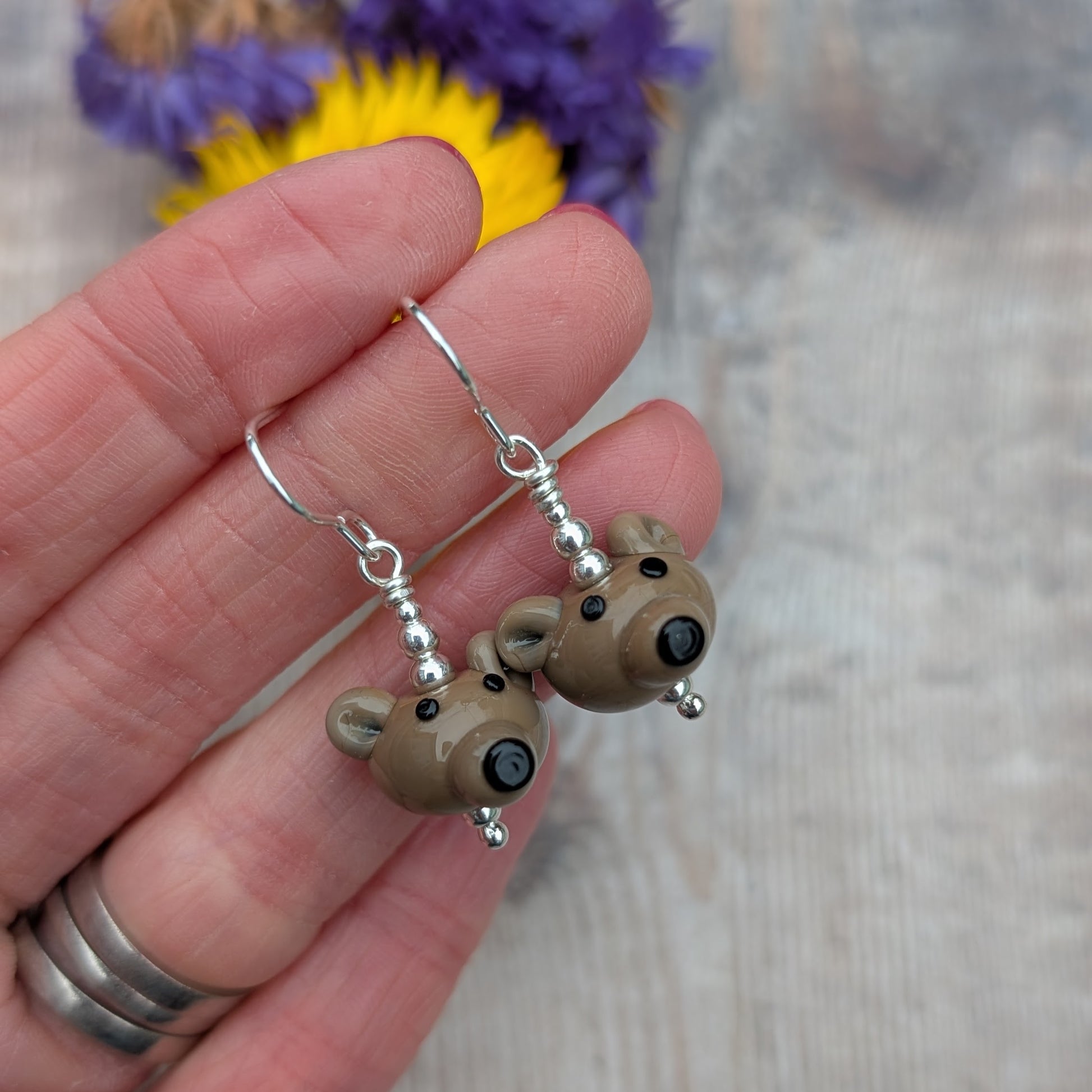 A hand holding the pair of bear earrings, highlighting the detailed glass bear faces and silver hooks.