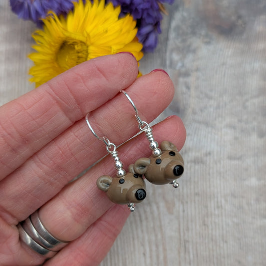 A hand holding the pair of bear earrings, highlighting the detailed glass bear faces and silver hooks.