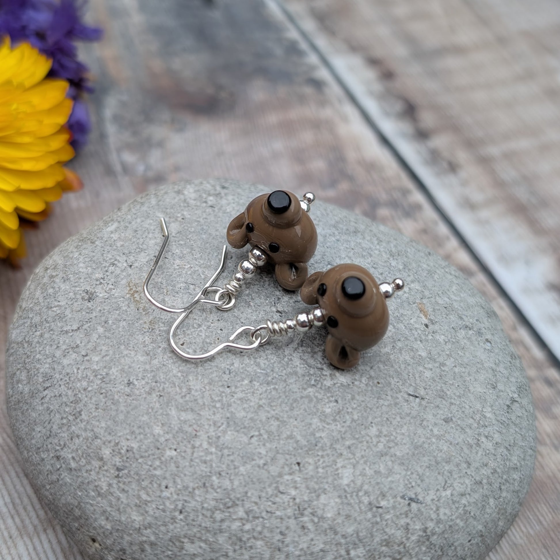 Close-up of silver earrings featuring handmade brown bear lampwork beads, resting on a smooth grey stone.