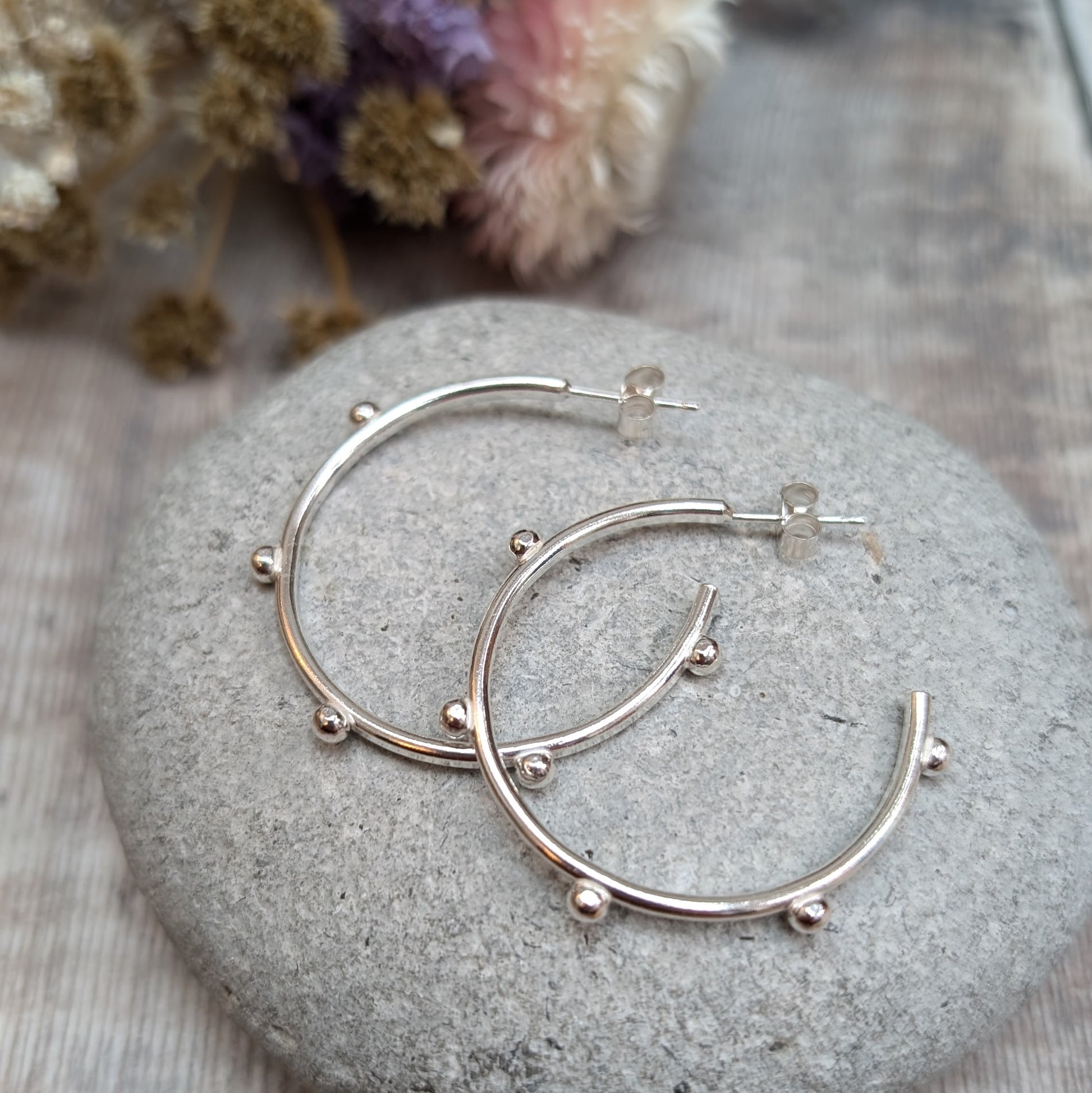 Pair of silver hoop earrings with small pebble-like details evenly spaced around the hoop, placed on a smooth stone with dried flowers in the background