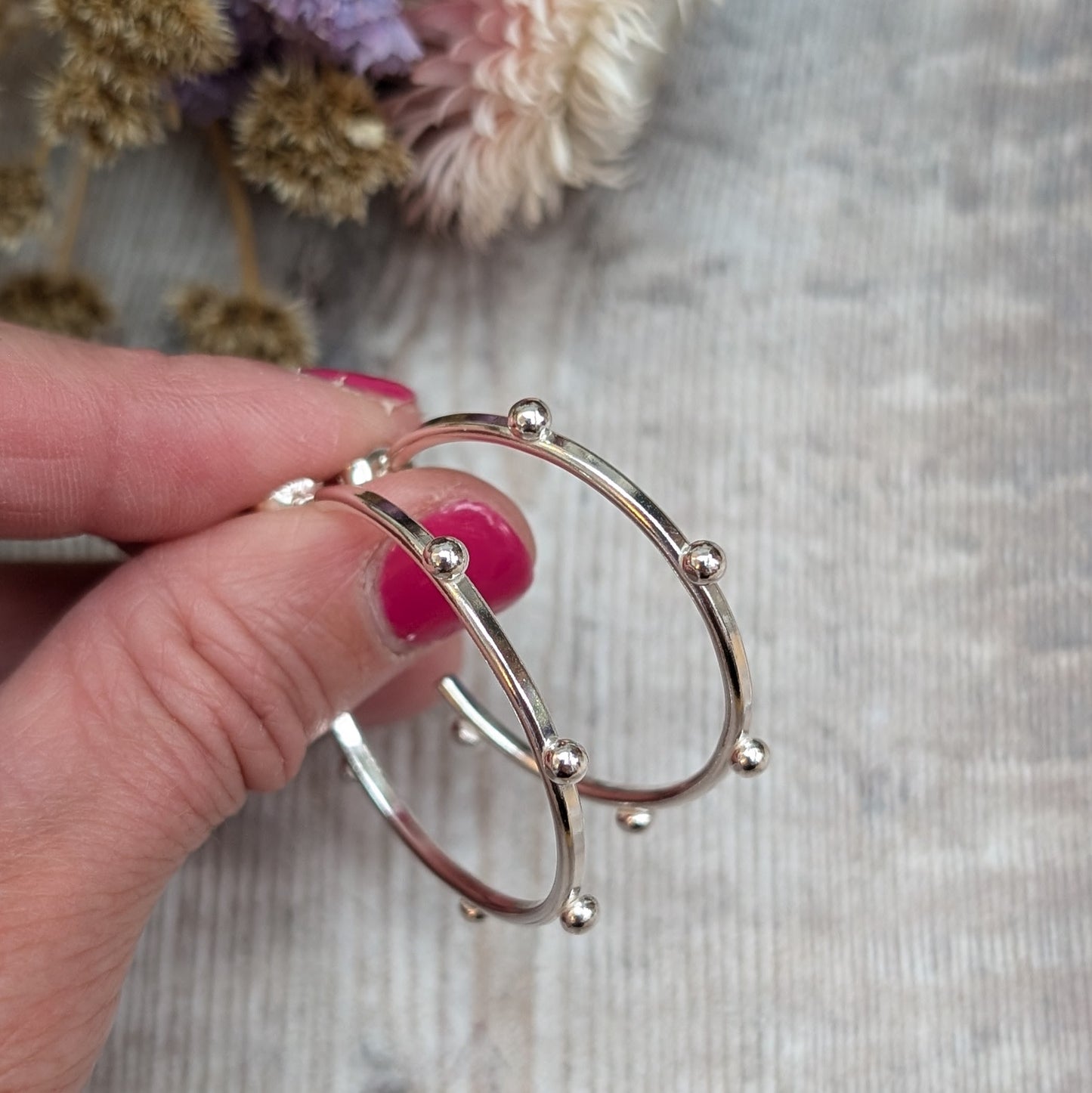 Close-up of a hand holding a pair of silver hoop earrings with small pebble-like details evenly spaced around the hoop, with dried flowers in the background.
