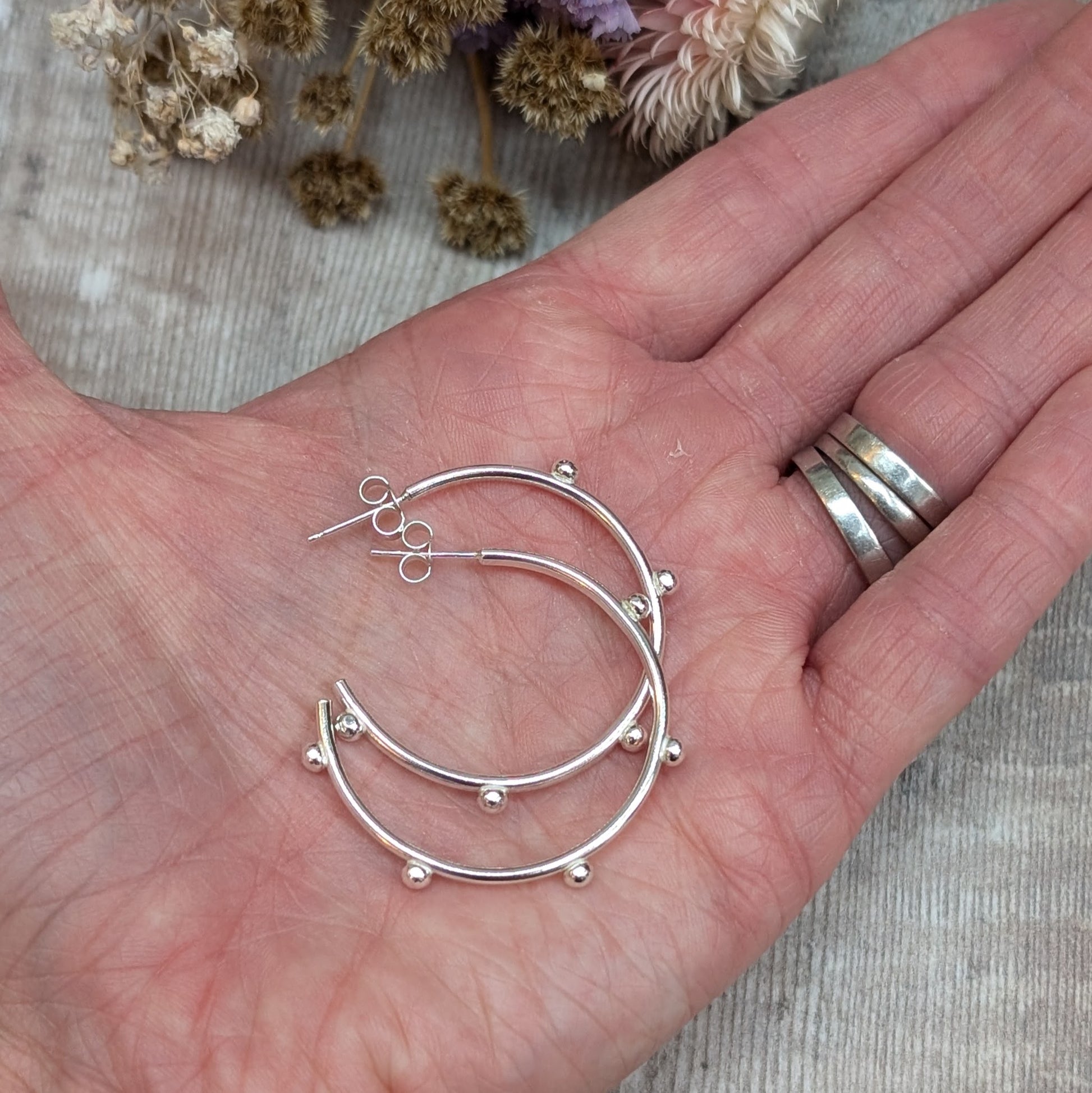 Close-up of a hand holding a pair of silver hoop earrings with small pebble-like details evenly spaced around the hoop, with dried flowers in the background.