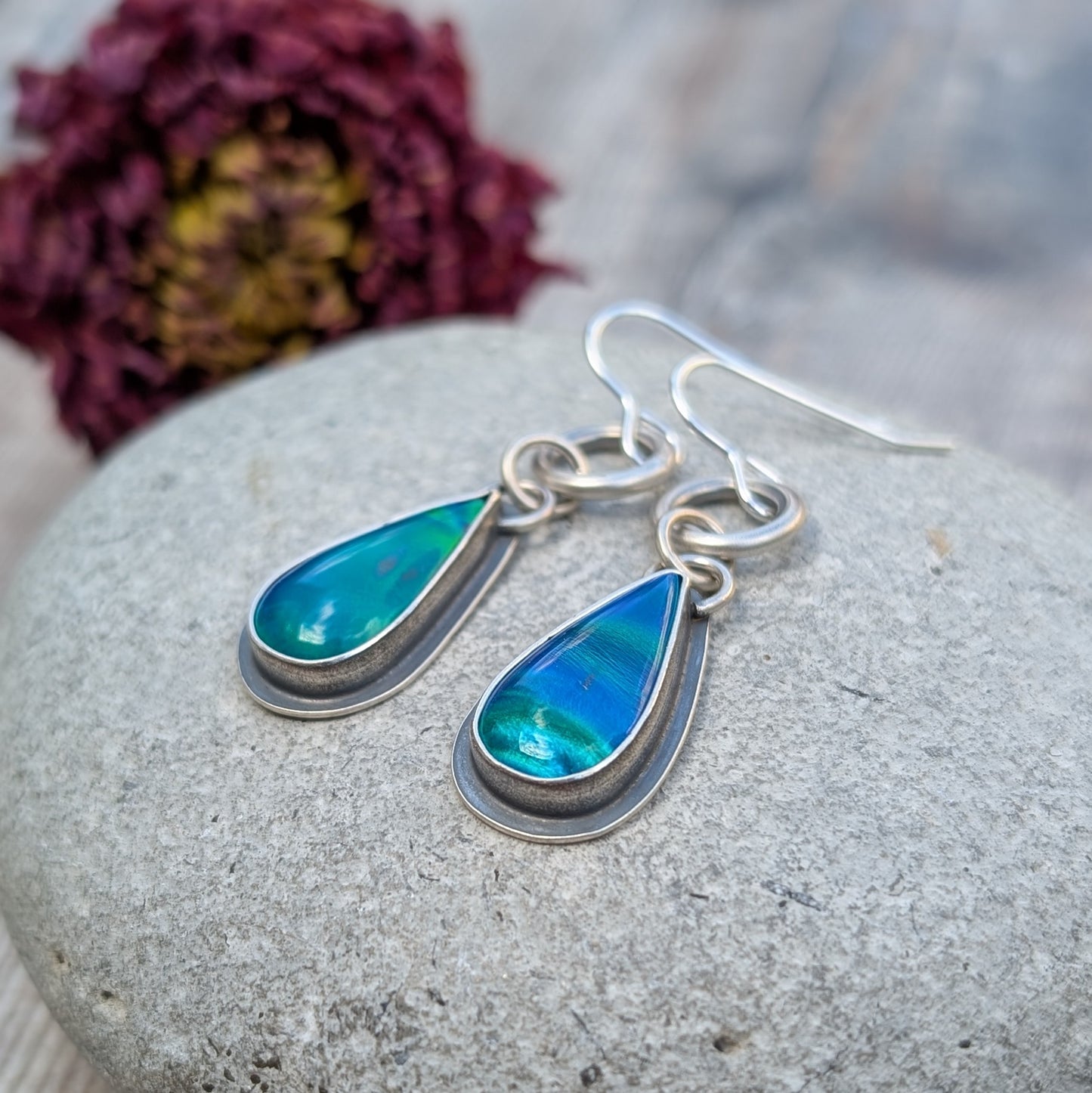 A pair of blue Aurora Opal teardrop earrings resting on a stone, capturing the depth and iridescence of the lab-grown opals. The oxidised silver setting adds contrast, highlighting the colours.