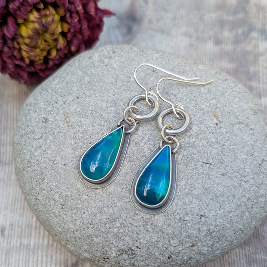 A pair of blue Aurora Opal teardrop earrings resting on a stone, capturing the depth and iridescence of the lab-grown opals. The oxidised silver setting adds contrast, highlighting the colours.