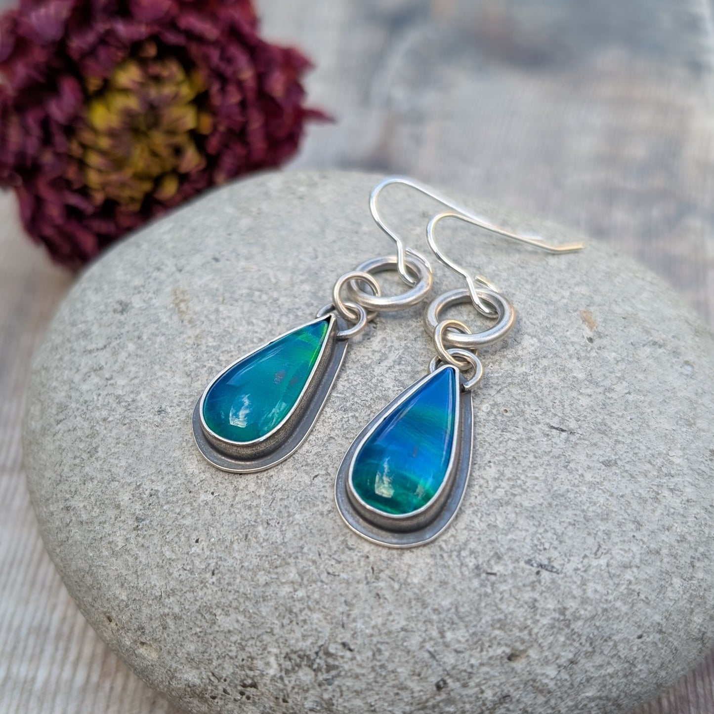 A pair of blue Aurora Opal teardrop earrings resting on a stone, capturing the depth and iridescence of the lab-grown opals. The oxidised silver setting adds contrast, highlighting the colours.