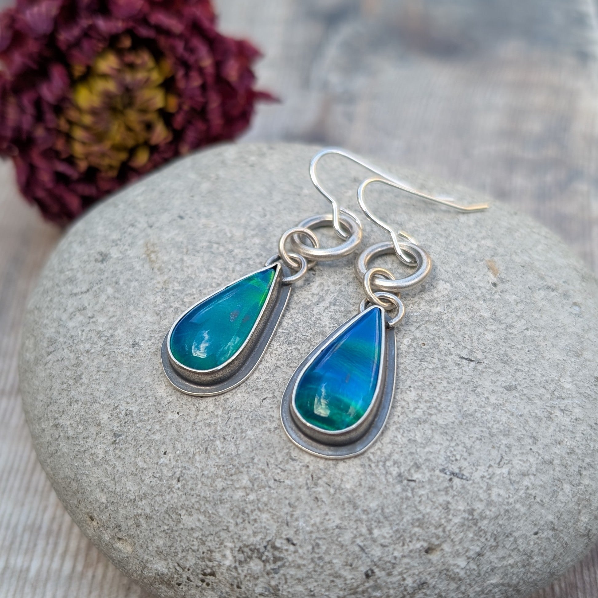 A pair of blue Aurora Opal teardrop earrings resting on a stone, capturing the depth and iridescence of the lab-grown opals. The oxidised silver setting adds contrast, highlighting the colours.