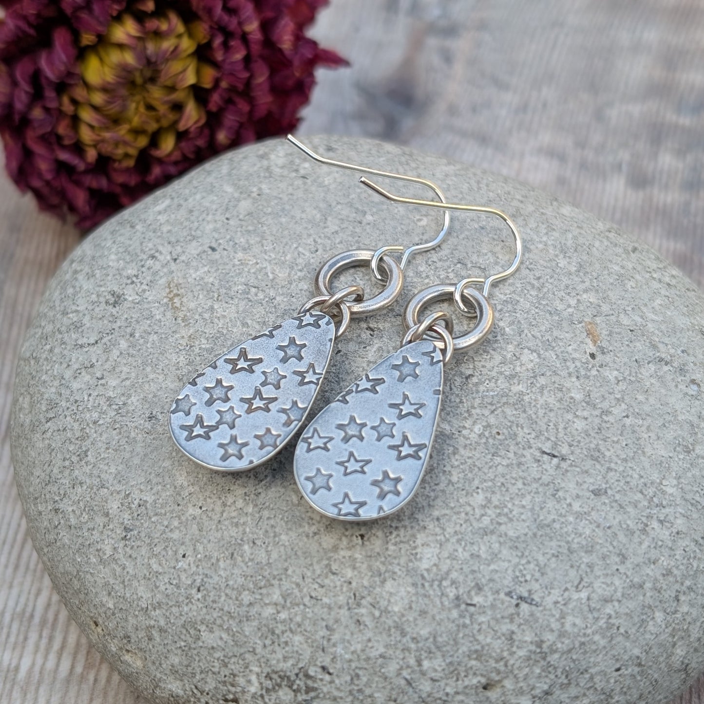 A pair of handmade sterling silver earrings with a hand-stamped star pattern on the back, resting on a smooth stone with a dried flower in the background. The oxidised finish enhances the celestial design.