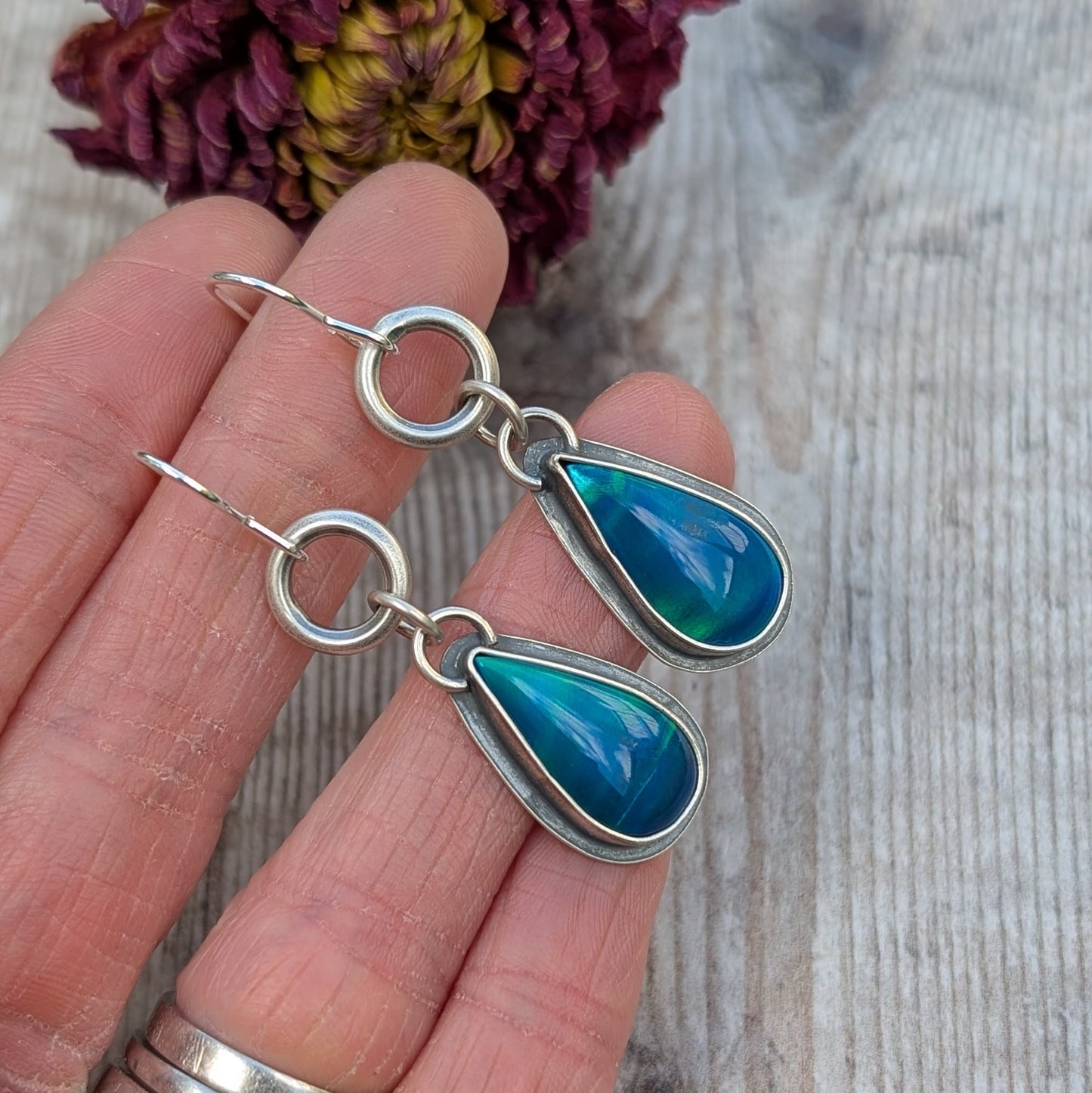 A close-up of a pair of blue Aurora Opal teardrop earrings set in recycled sterling silver, held in a person’s hand. The opals shimmer with shades of blue and green, framed by oxidised silver.