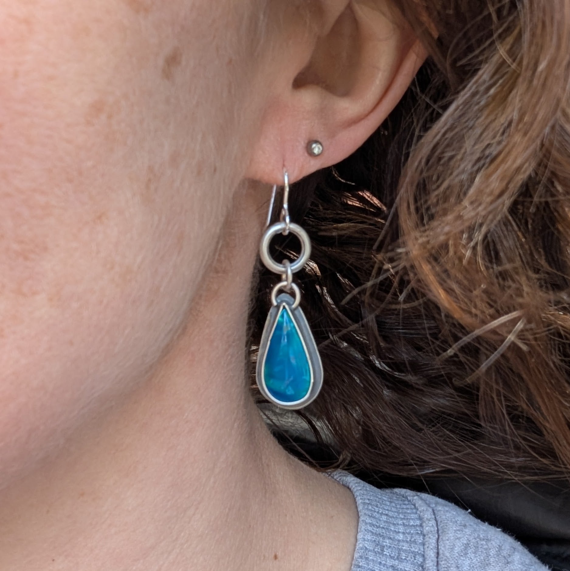 One earring from the blue Aurora Opal teardrop earrings worn on a person’s ear, showcasing its size, movement, and vibrant play of colours. The handmade silver links add a touch of elegance.