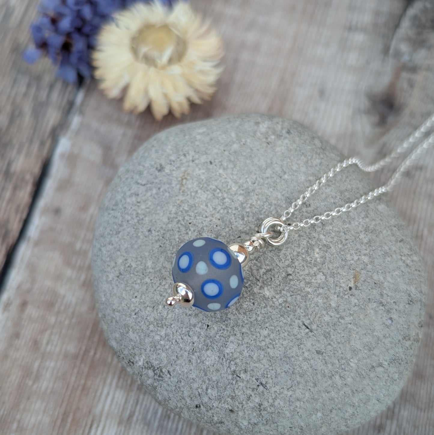 Blue Spotty Lampwork Glass Necklace resting on a smooth stone with dried flowers in the background.