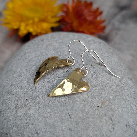 A pair of brass hammered curved heart earrings resting on a smooth grey stone with yellow and orange dried flowers in the background.