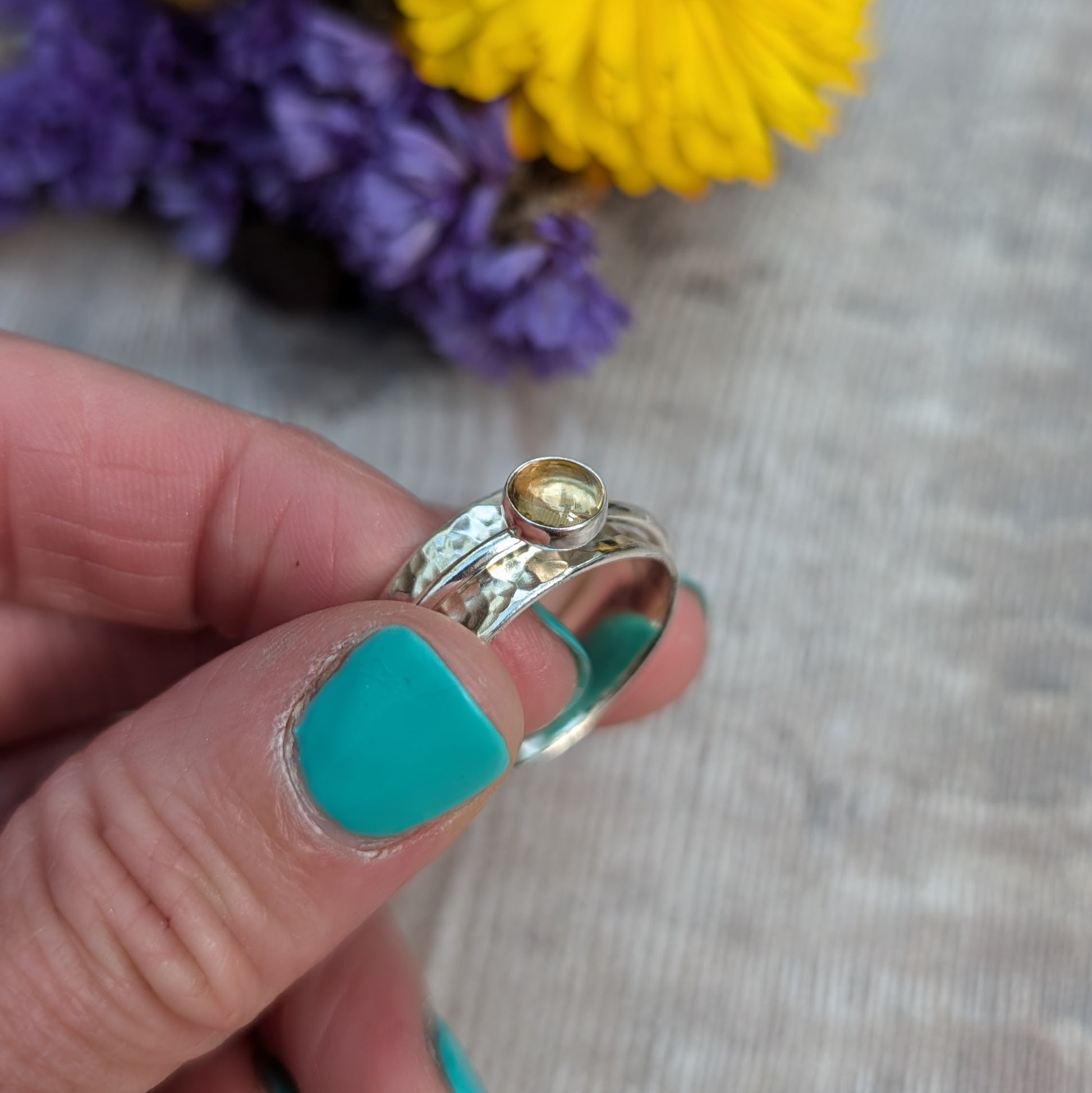 A close-up of a handmade citrine spinner ring held between fingers, highlighting the hammered sterling silver band and the golden citrine gemstone. The background features soft-focus yellow and purple flowers.