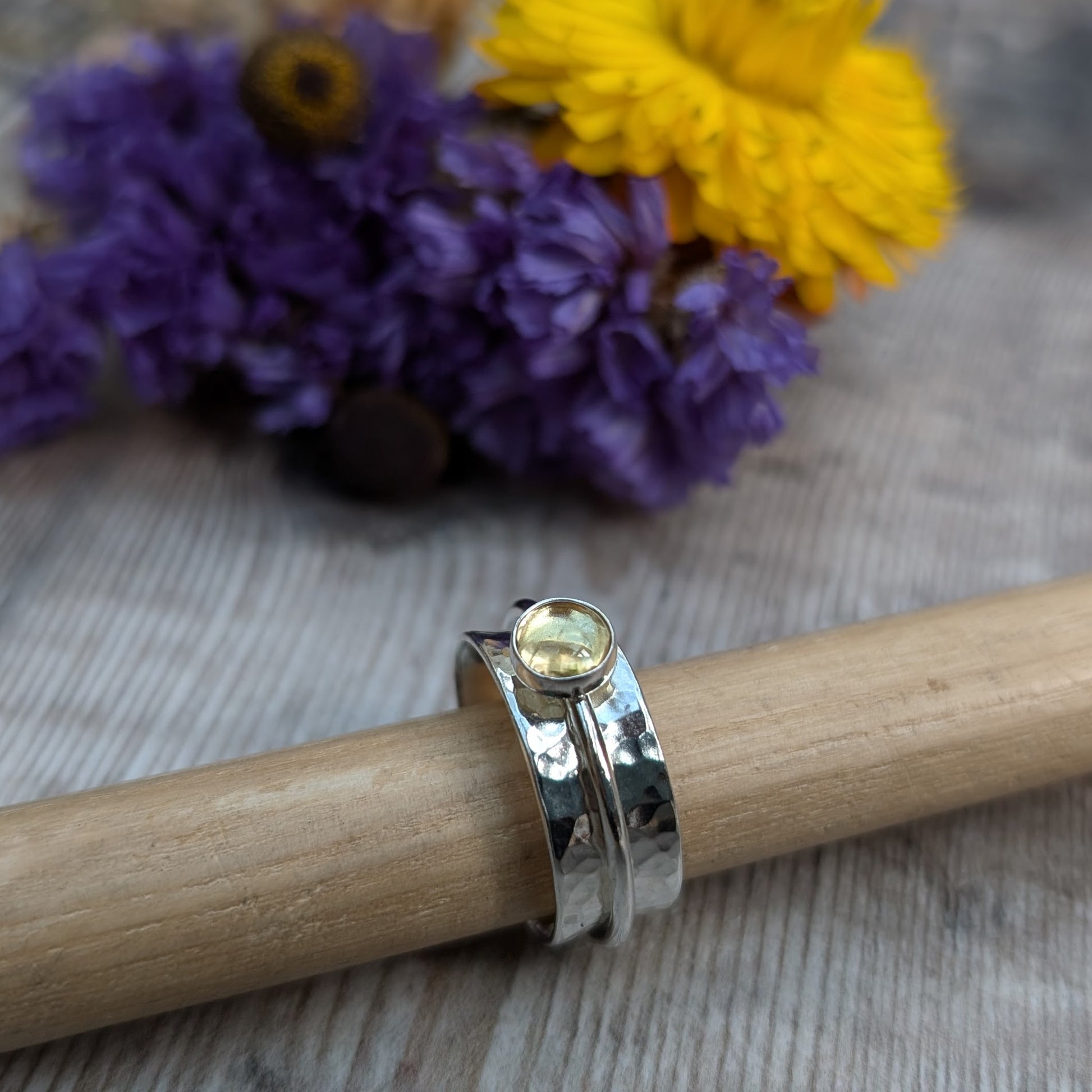A close-up of a handmade citrine spinner ring resting on a wooden rod, featuring a recycled sterling silver band with a hammered texture and a warm yellow citrine gemstone in a bezel setting. The background includes vibrant yellow and purple flowers.