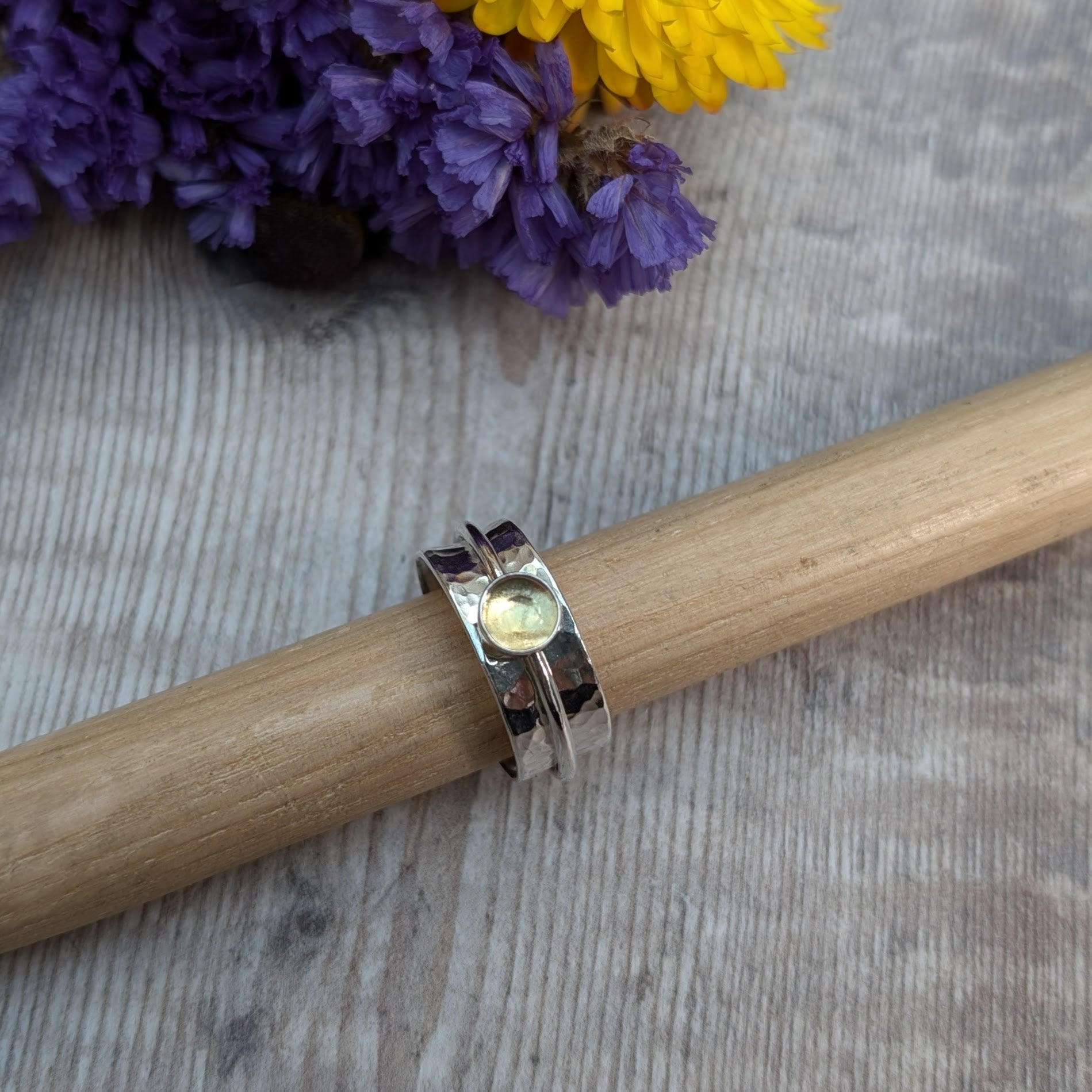 A close-up of a handmade citrine spinner ring resting on a wooden rod, featuring a recycled sterling silver band with a hammered texture and a warm yellow citrine gemstone in a bezel setting. The background includes vibrant yellow and purple flowers.