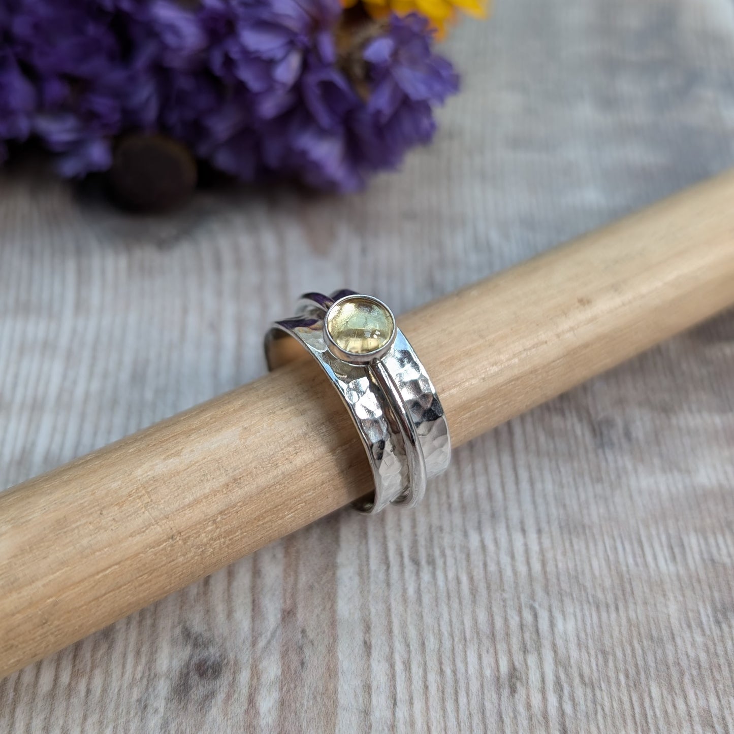 A close-up of a handmade citrine spinner ring resting on a wooden rod, featuring a recycled sterling silver band with a hammered texture and a warm yellow citrine gemstone in a bezel setting. The background includes vibrant yellow and purple flowers.