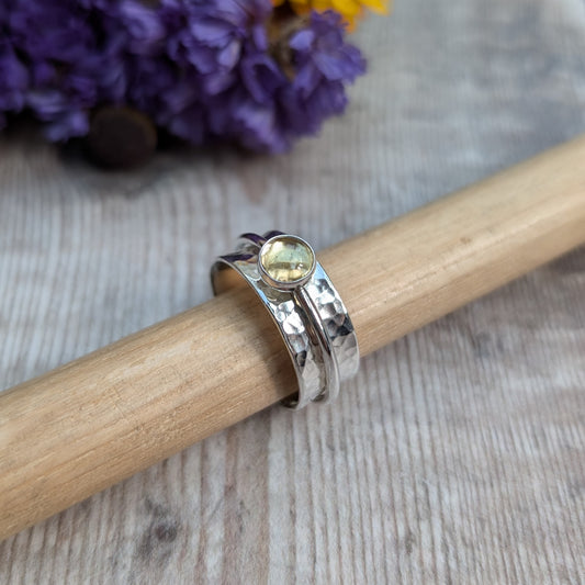 A close-up of a handmade citrine spinner ring resting on a wooden rod, featuring a recycled sterling silver band with a hammered texture and a warm yellow citrine gemstone in a bezel setting. The background includes vibrant yellow and purple flowers.