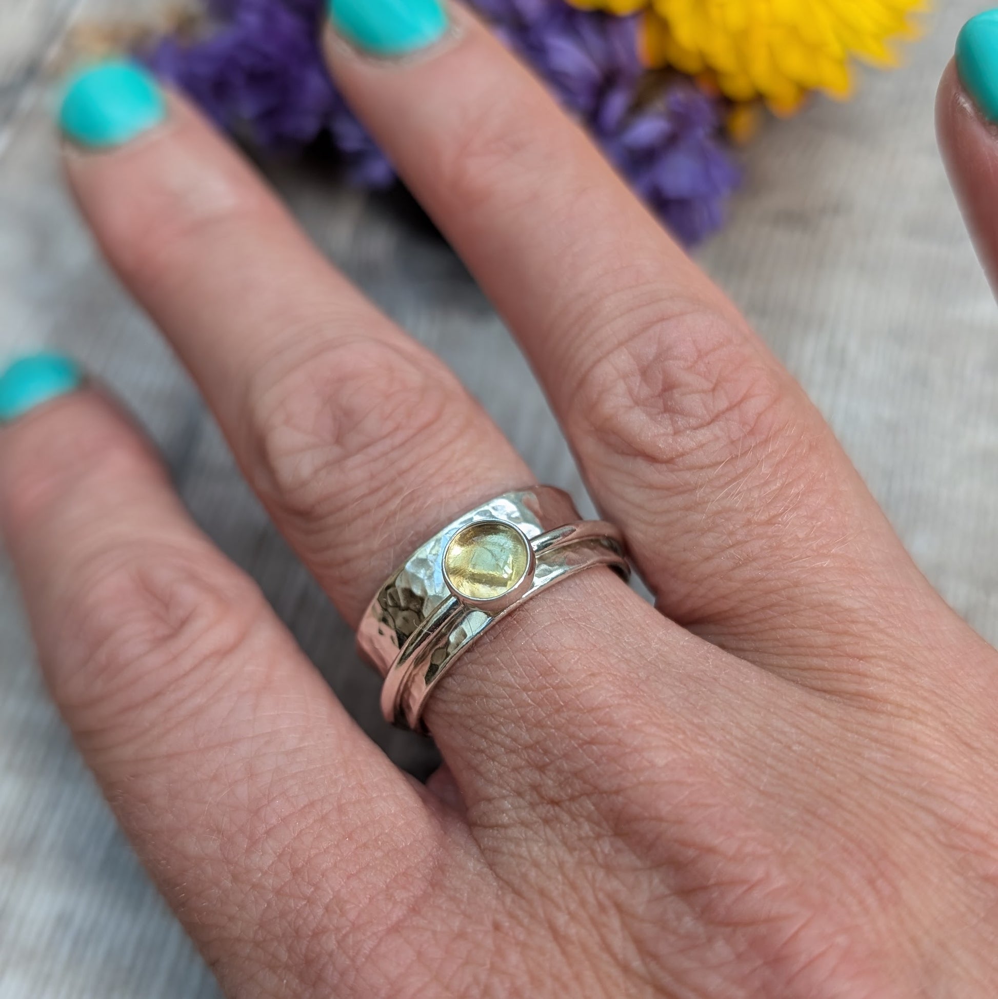 A handmade citrine spinner ring worn on a hand, showcasing its textured sterling silver band and a bezel-set citrine stone. The hand has turquoise nail polish, and blurred flowers in the background add a natural touch.